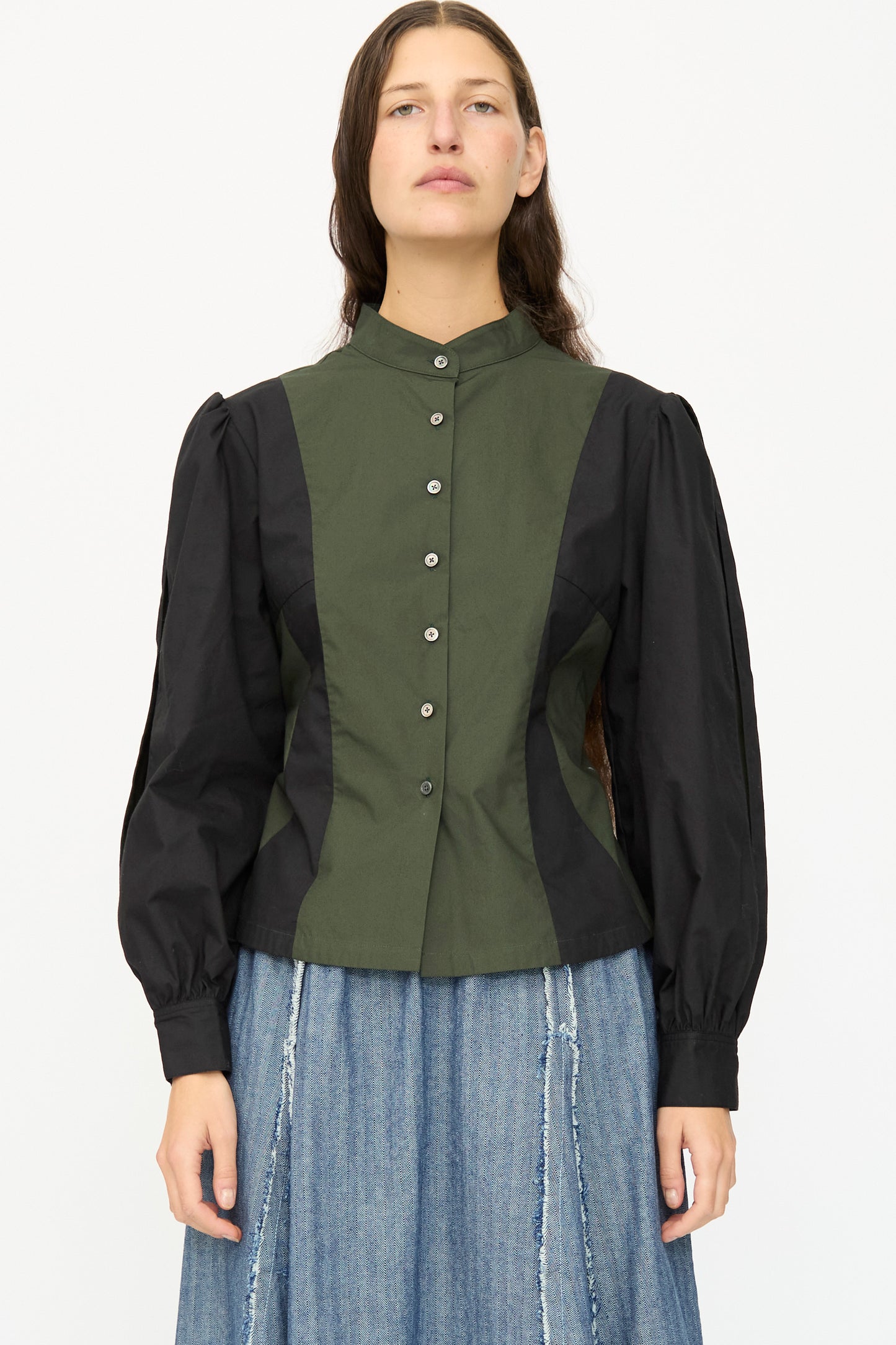 A person wears the SC103 Poplin Folio Blouse in Bolt—a color blocked cotton poplin top with bishop sleeves, high collar, and button front—paired with a blue denim skirt.