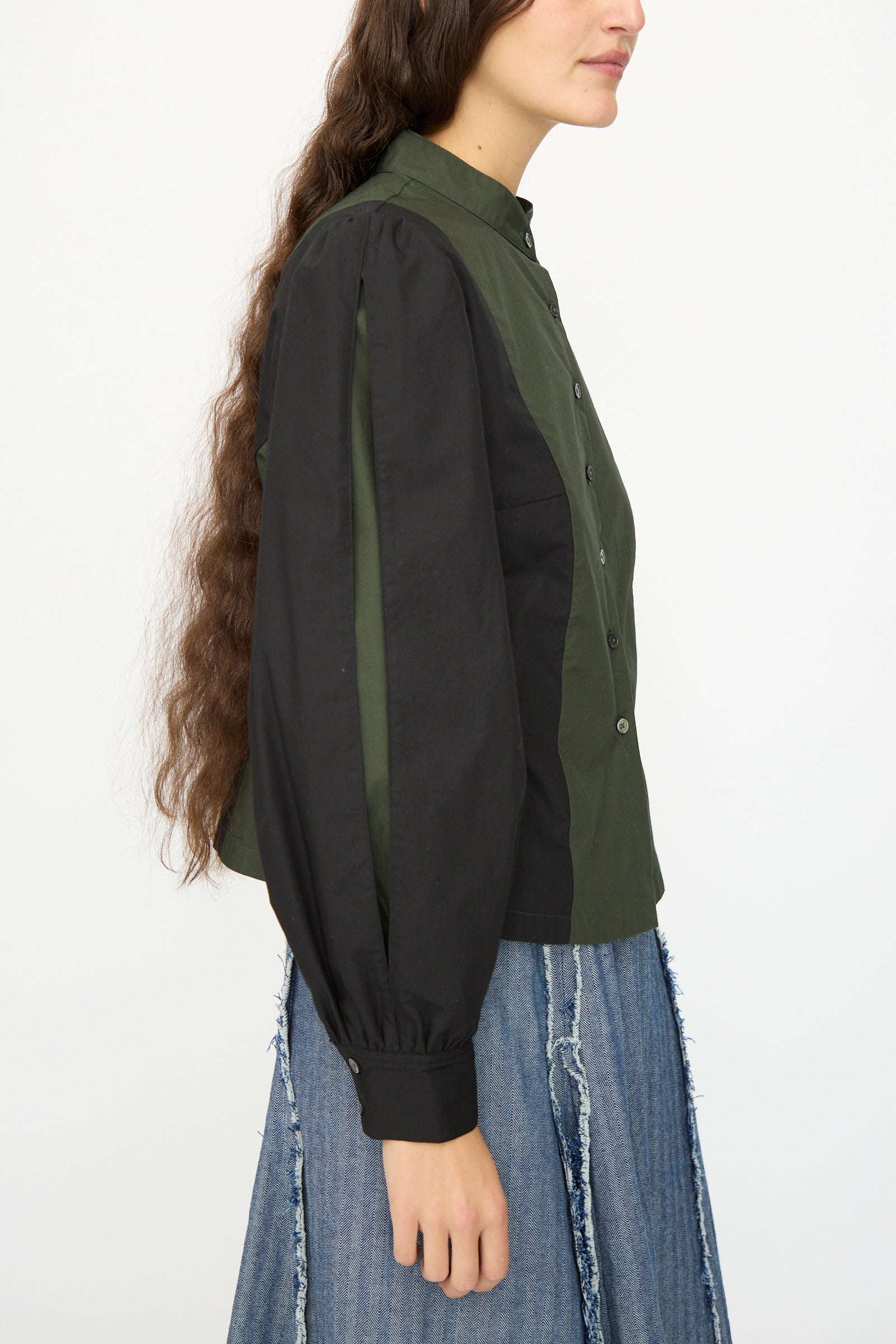 A woman with long brown hair is shown from the side wearing the SC103 Poplin Folio Blouse in Bolt, a green color-blocked cotton poplin button-up with black sleeves, paired with a frayed denim skirt.