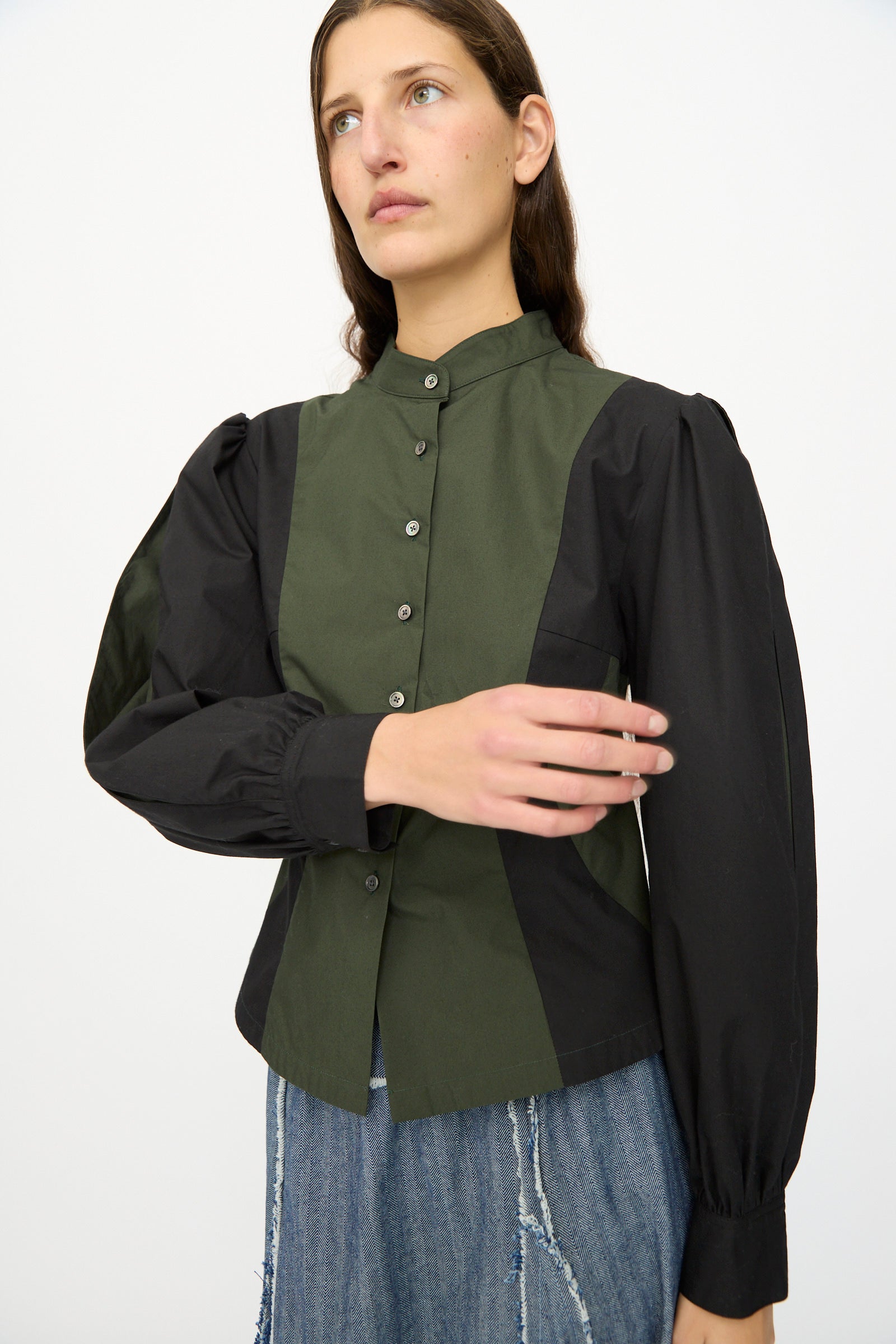 A person wears the SC103 Poplin Folio Blouse in Bolt—a colorblocked cotton poplin top with black bishop sleeves and olive green front panels—paired with blue jeans, standing against a plain white background.