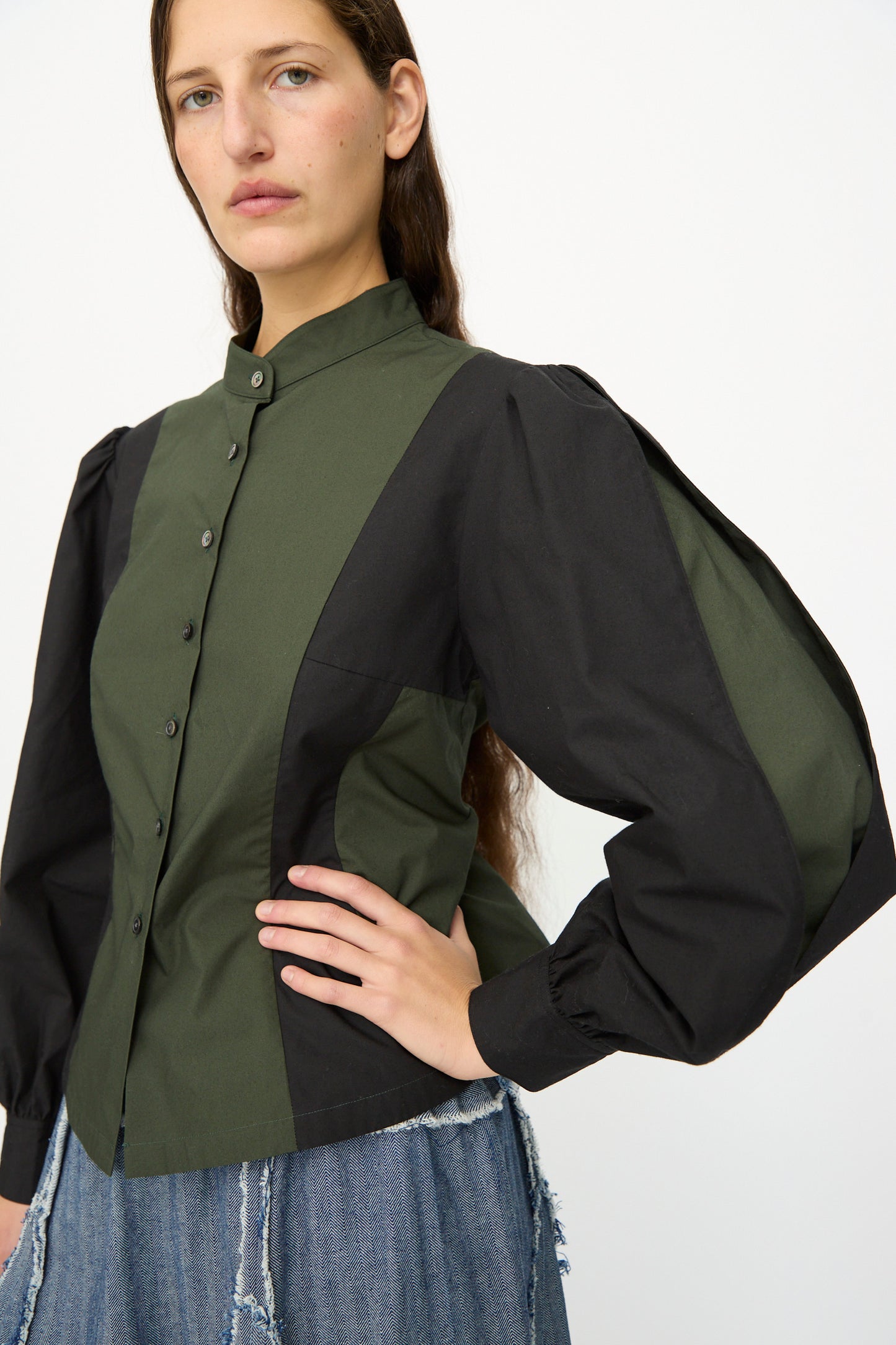 A woman stands against a white background wearing the SC103 Poplin Folio Blouse in Bolt—an olive green and black color-blocked cotton poplin blouse with long bishop sleeves—paired with denim jeans.