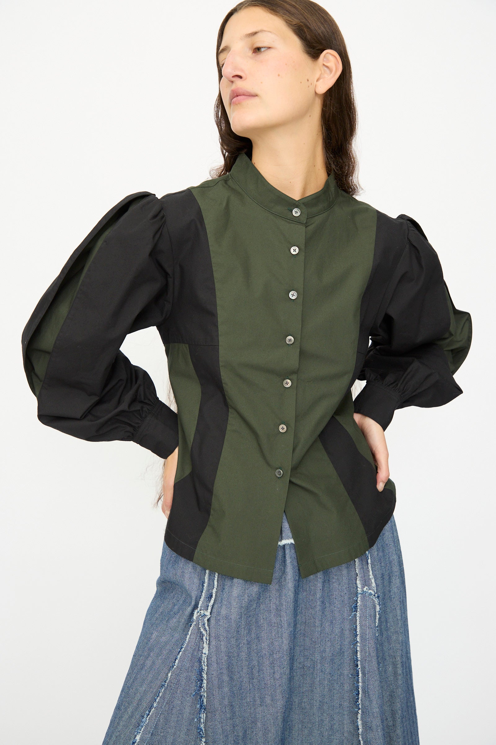 A woman in the SC103 Poplin Folio Blouse in Bolt—green and black color-blocked poplin with bishop sleeves—and blue wide-leg jeans stands with one hand on her hip against a plain background.