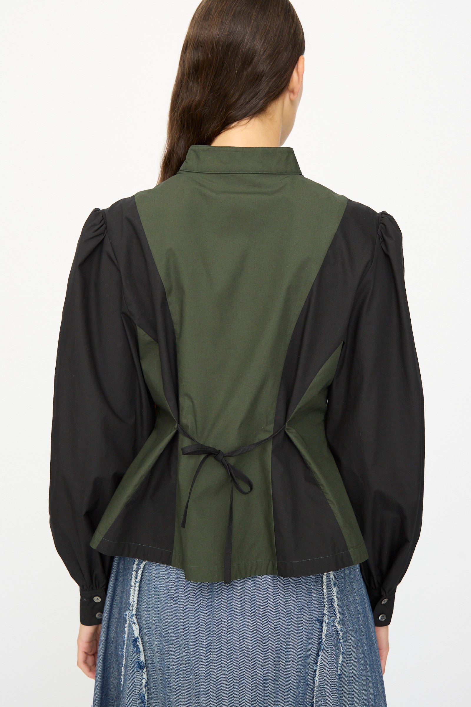 A woman with long brown hair is shown from the back wearing the SC103 Poplin Folio Blouse in Bolt, featuring color-blocked green and black cotton poplin, bishop sleeves, a back tie, and a distressed denim skirt.