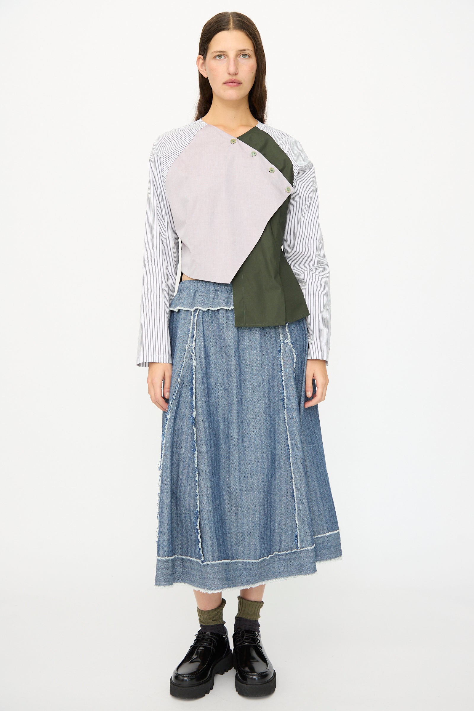 A woman faces forward in the SC103 Poplin Mix Peak Blouse in Horizon, a raw-edge denim skirt, green socks, and black platform shoes against a plain white background.