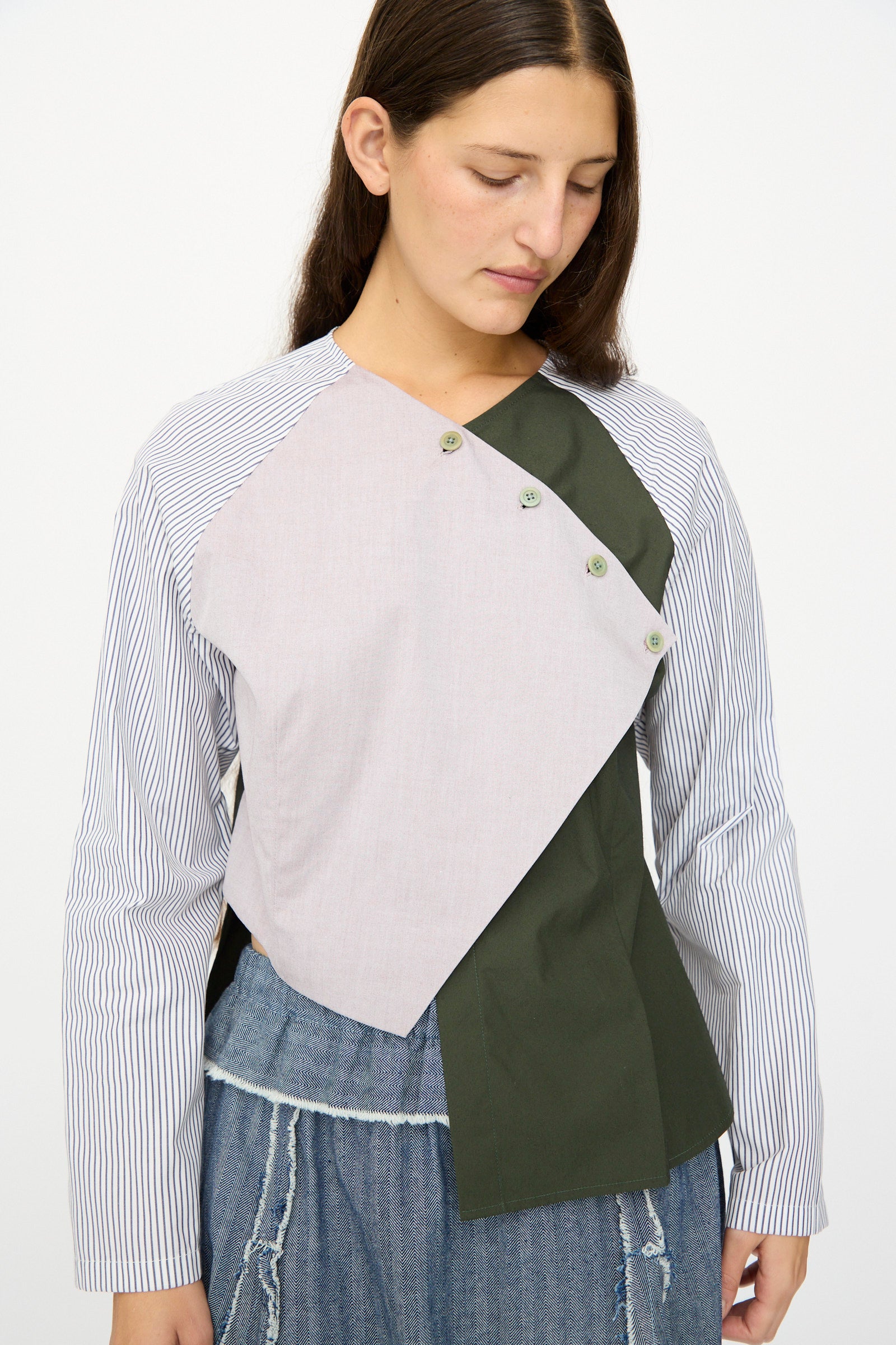 A woman with long brown hair models the SC103 Poplin Mix Peak Blouse in Horizon, featuring color-blocked, asymmetrical cotton poplin, paired with blue frayed jeans against a plain white background.