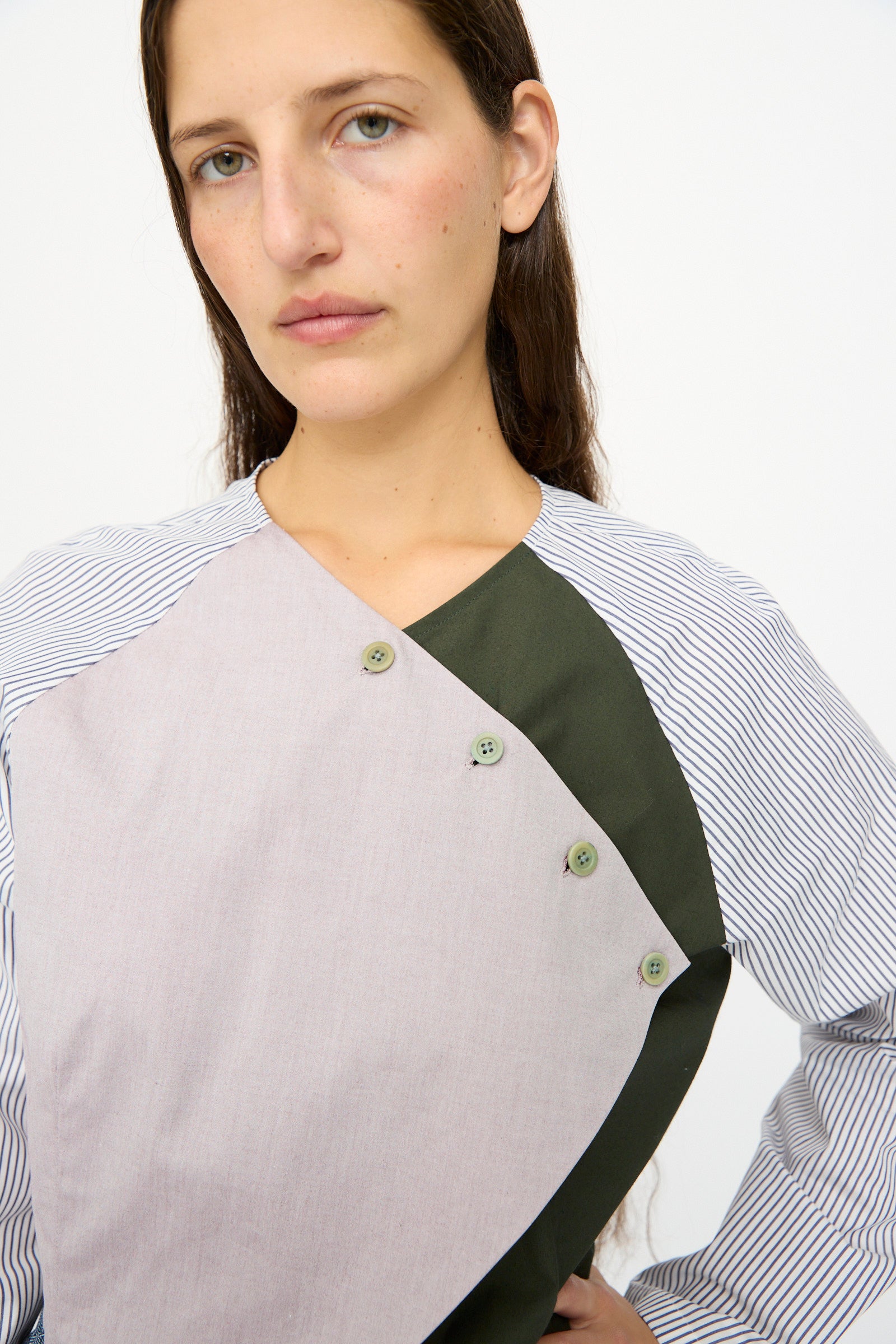 A woman with long brown hair wears the SC103 Poplin Mix Peak Blouse in Horizon, an asymmetrical cotton top with striped, beige, and green panels and four chest buttons, made in NYC. She stands against a plain white background.