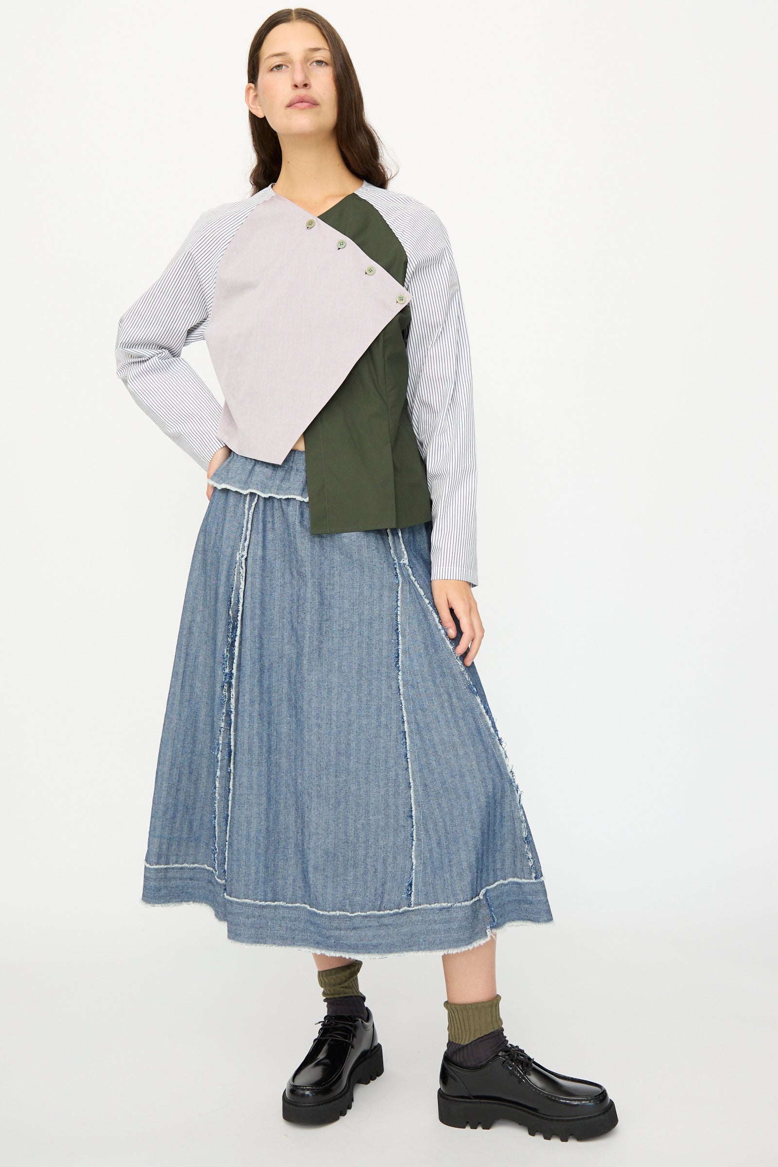 A woman stands against a plain white background wearing SC103’s Poplin Mix Peak Blouse in Horizon, an asymmetrical cotton blouse with button details, styled with a long denim skirt, socks, and black platform shoes. Made in NYC.
