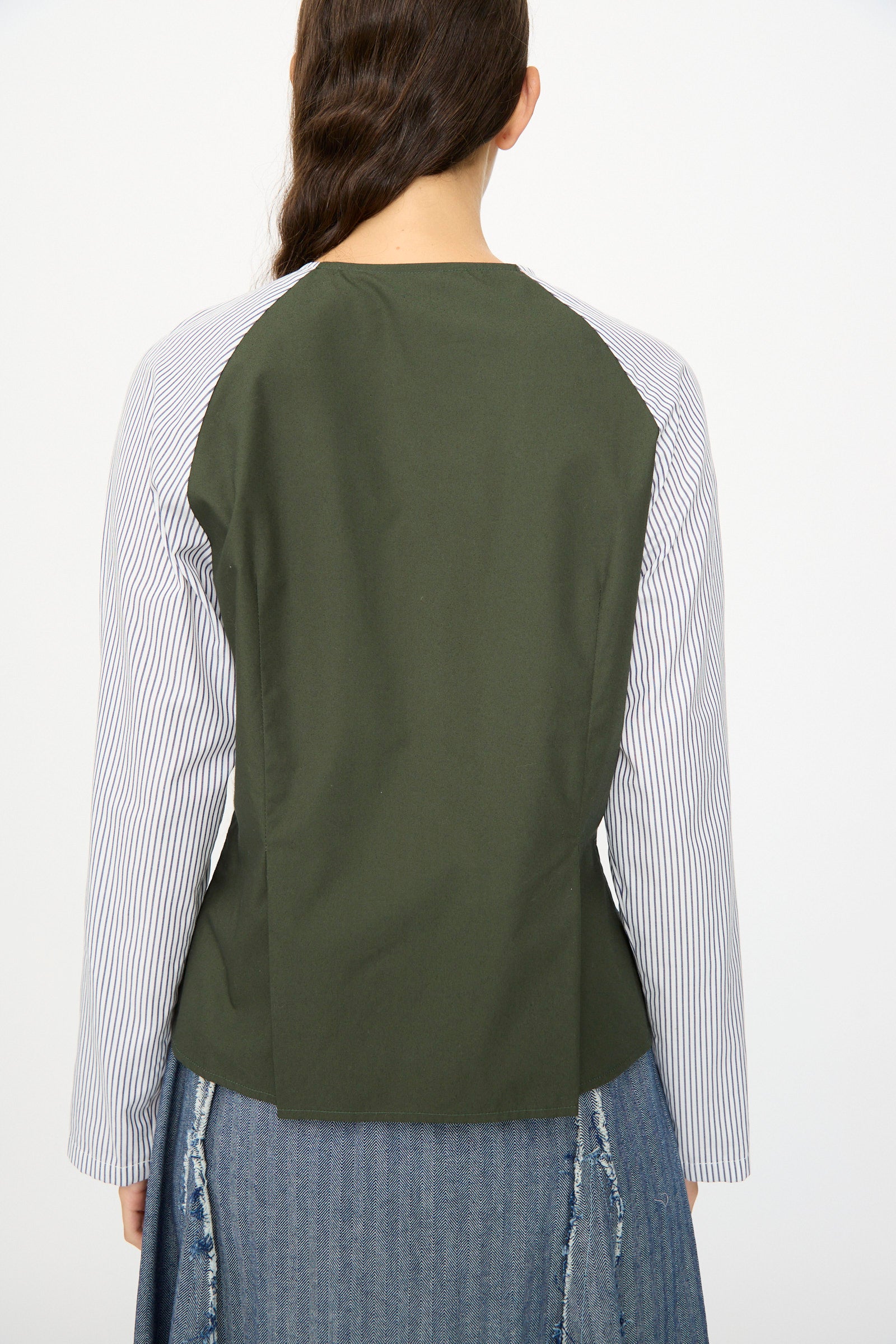 A person faces away, wearing the SC103 Poplin Mix Peak Blouse in Horizon—featuring striped white sleeves and a solid dark green back—paired with a blue denim skirt.
