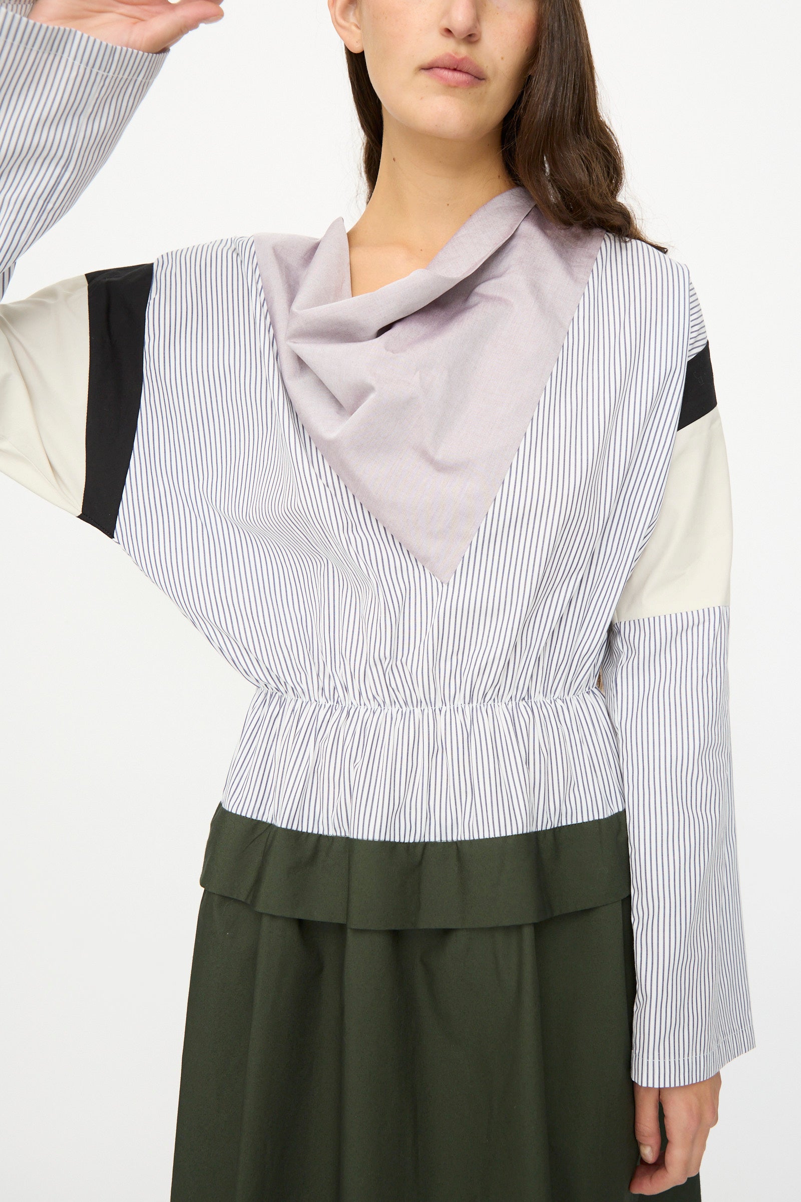 A person wearing the SC103 Poplin Mix Solace Dress in Horizon, styled with a dark green skirt and cowl neck, raises their left arm. The long-sleeved, multi-patterned dress partially obscures the face.
