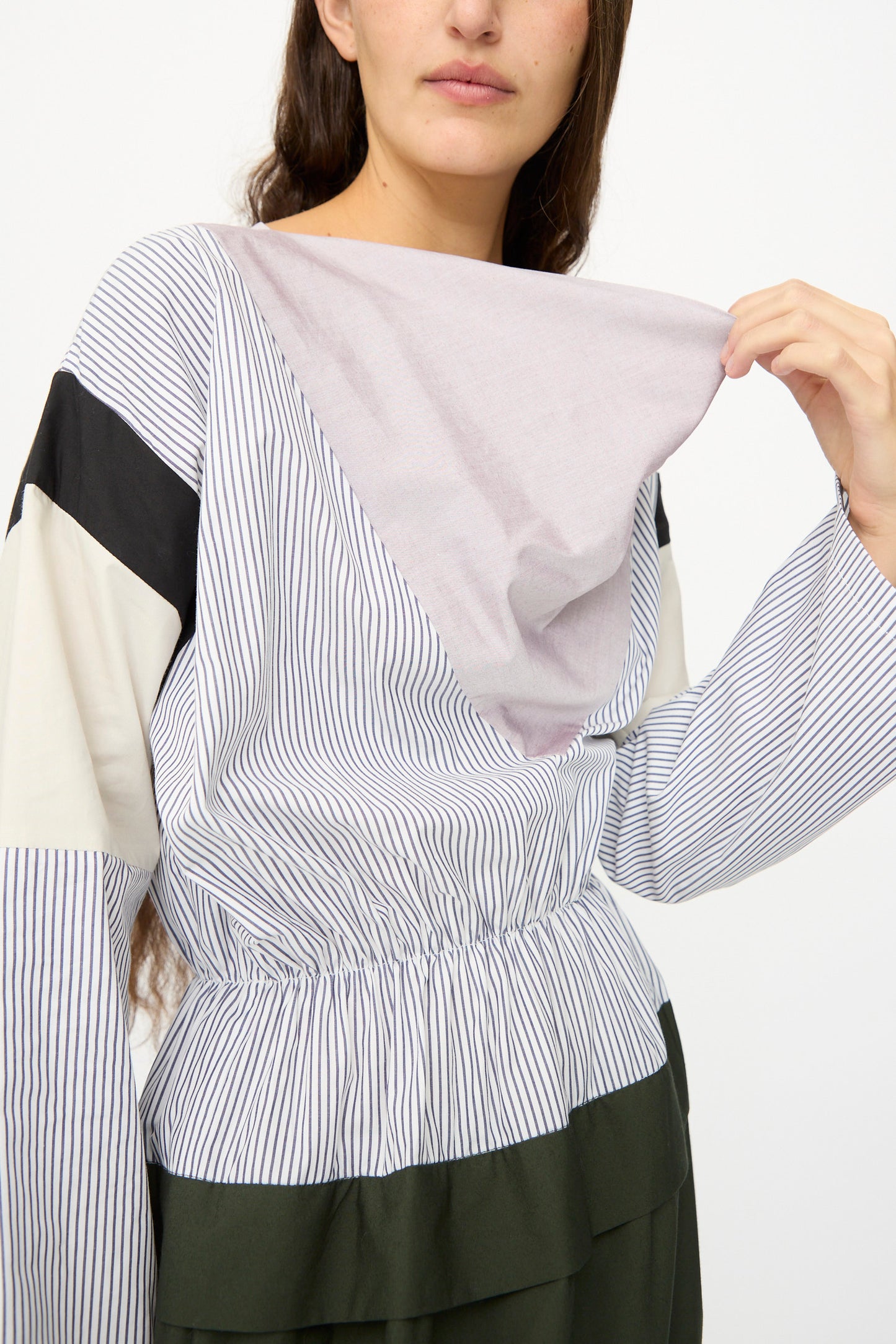 A person wearing the SC103 Poplin Mix Solace Dress in Horizon, a long sleeve dress with multi-patterned stripes and color blocks made in NYC, pulls at the fabric near the neckline.