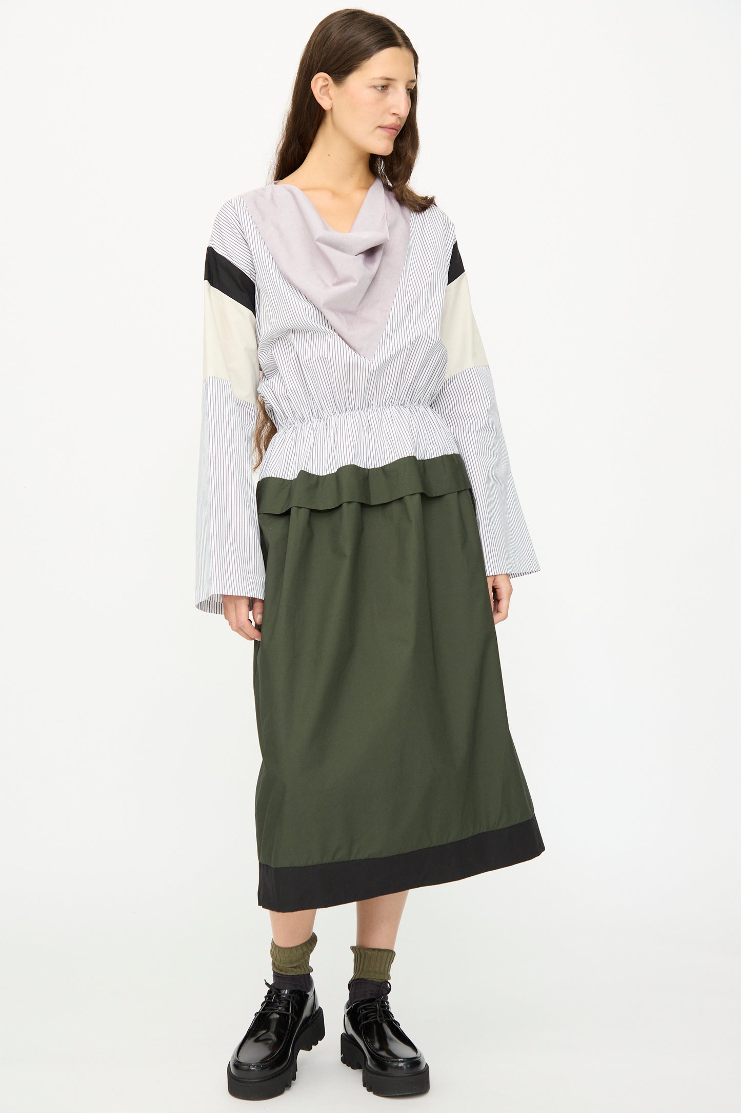 A woman stands against a white background, wearing the SC103 Poplin Mix Solace Dress in Horizon, featuring long sleeves, a color-blocked draped collar, and paired with black platform shoes and socks.