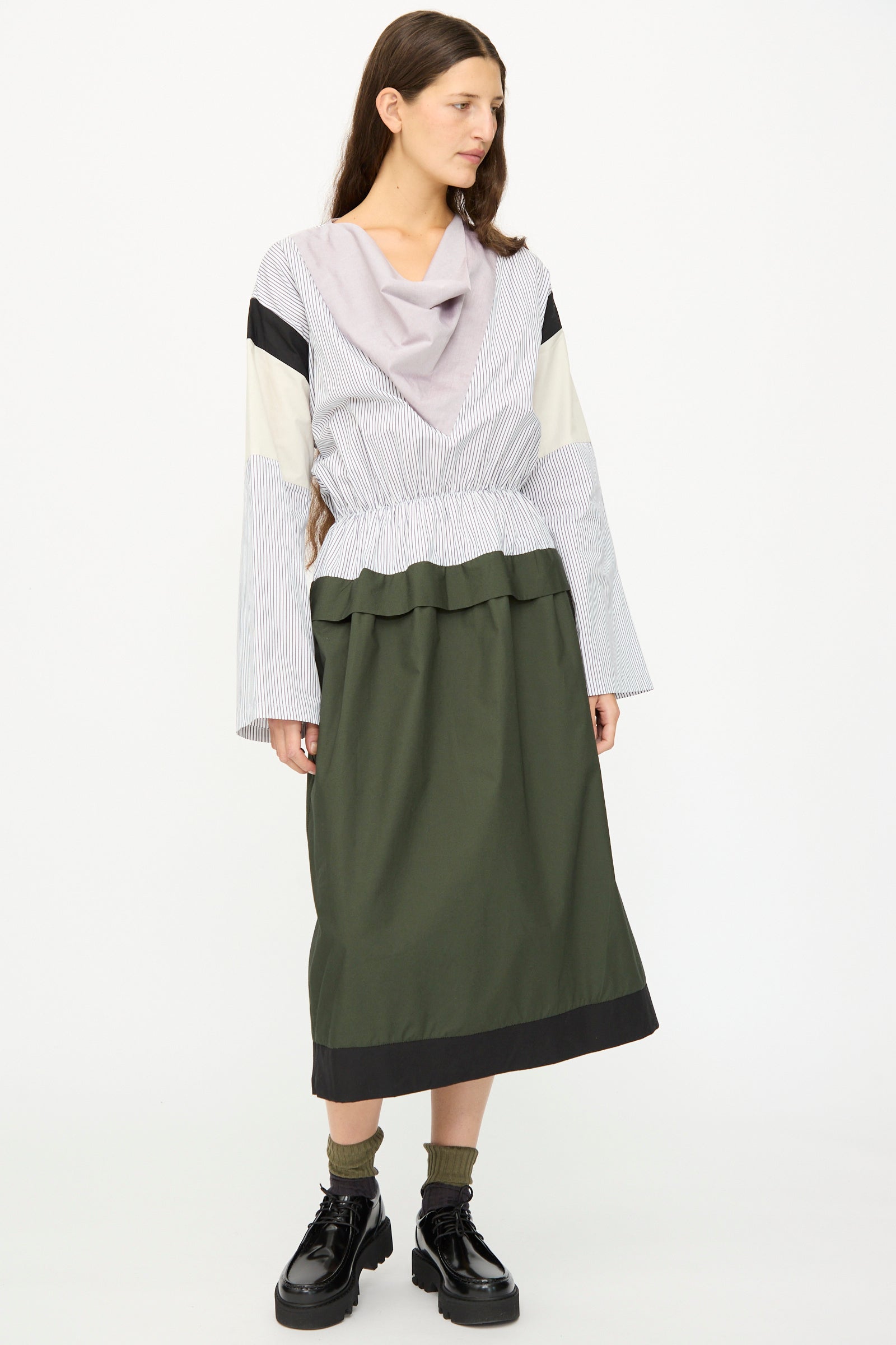 A woman stands against a white background, wearing the SC103 Poplin Mix Solace Dress in Horizon, featuring long sleeves, a color-blocked draped collar, and paired with black platform shoes and socks.