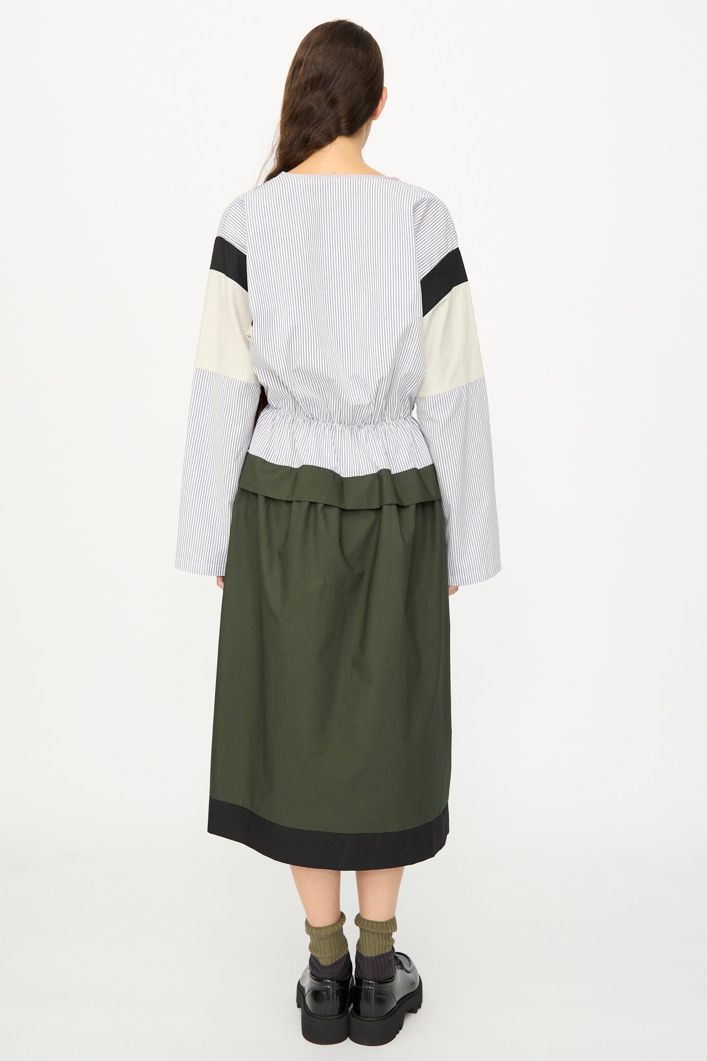 A person faces away, wearing the SC103 Poplin Mix Solace Dress in Horizon—a color-blocked, long sleeve style with stripes and a green cotton poplin midi skirt—paired with black shoes and green socks; made in NYC.