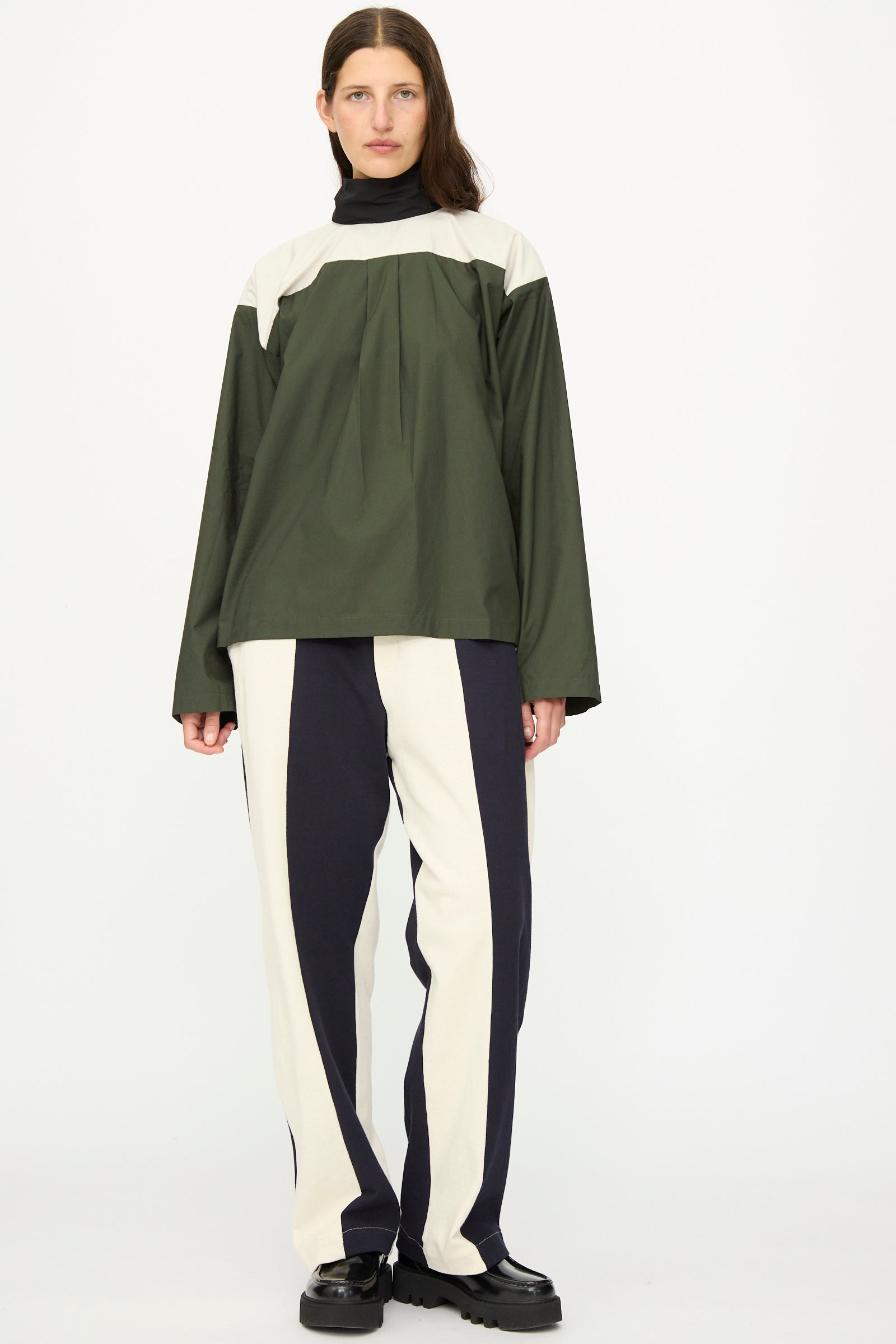 A person in SC103 Rib Knit Leap Sweats in Index stands against a plain white background, wearing relaxed two-tone cream and black pants, a loose olive green and beige long-sleeve top, black turtleneck, and black shoes.