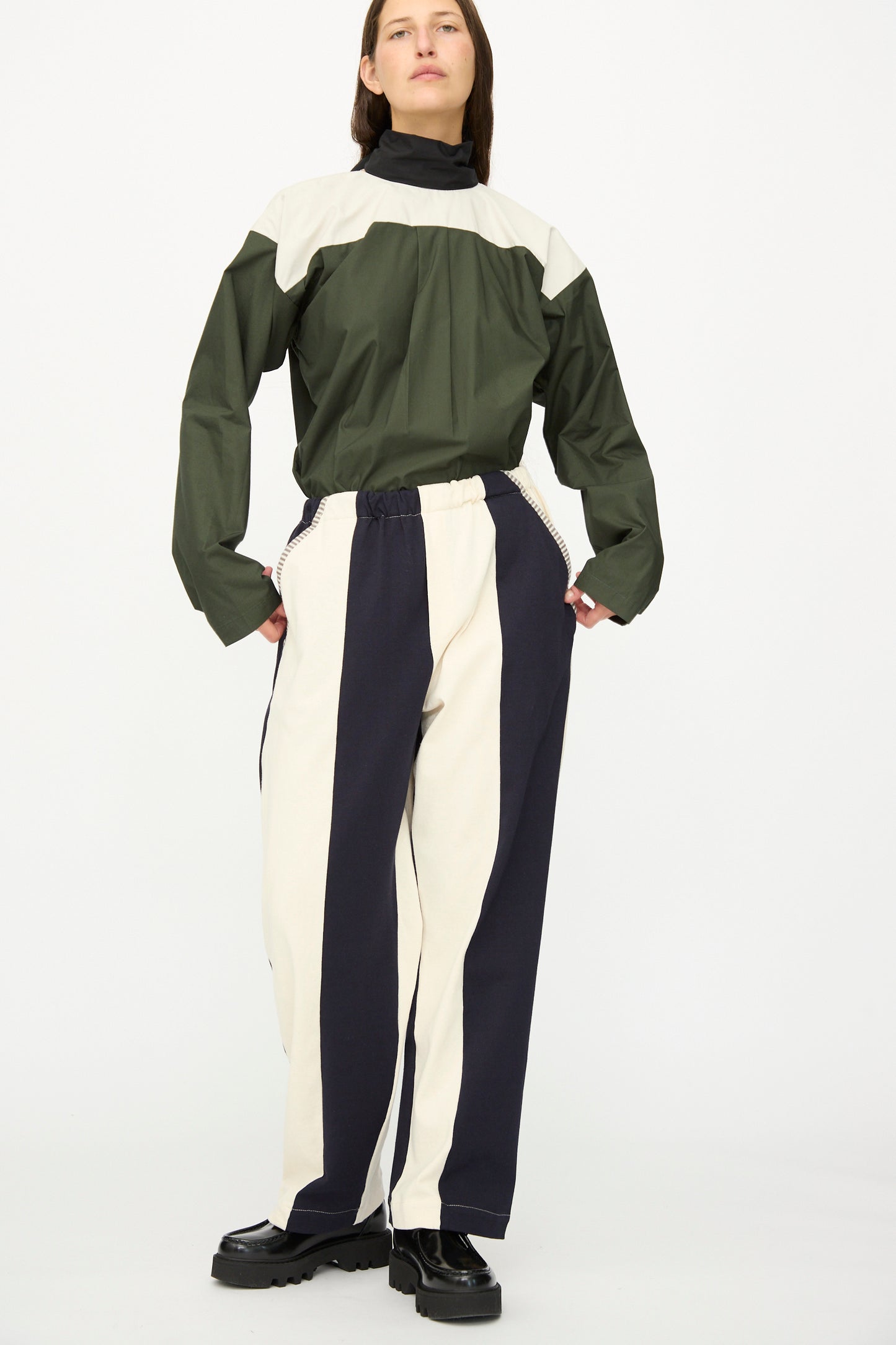 A person wears SC103 Rib Knit Leap Sweats in Index—black and white striped cotton knit pants with a relaxed fit—paired with a green and beige top and black shoes against a plain background. The stylish pants are made in NYC.