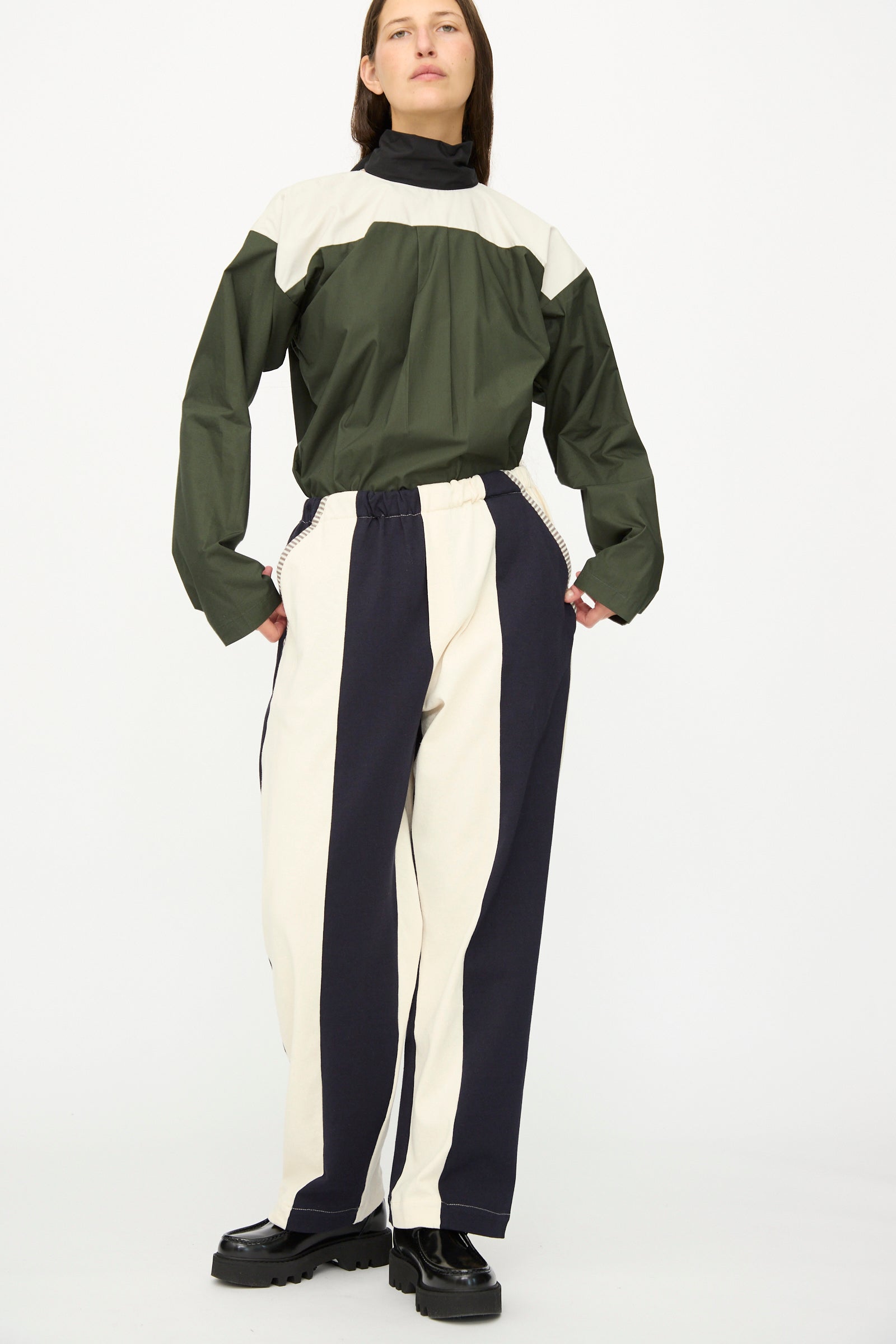 A person wears SC103 Rib Knit Leap Sweats in Index—black and white striped cotton knit pants with a relaxed fit—paired with a green and beige top and black shoes against a plain background. The stylish pants are made in NYC.