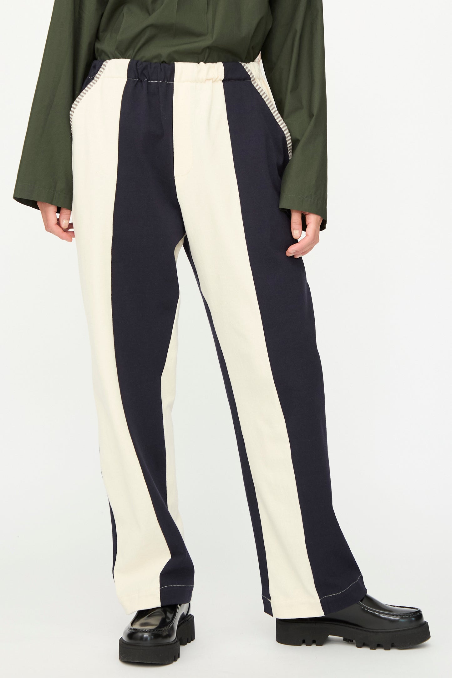 Wearing SC103's Rib Knit Leap Sweats in Index—black and white striped knit pants made in NYC—with a green long-sleeve top and black platform shoes, standing against a plain background.