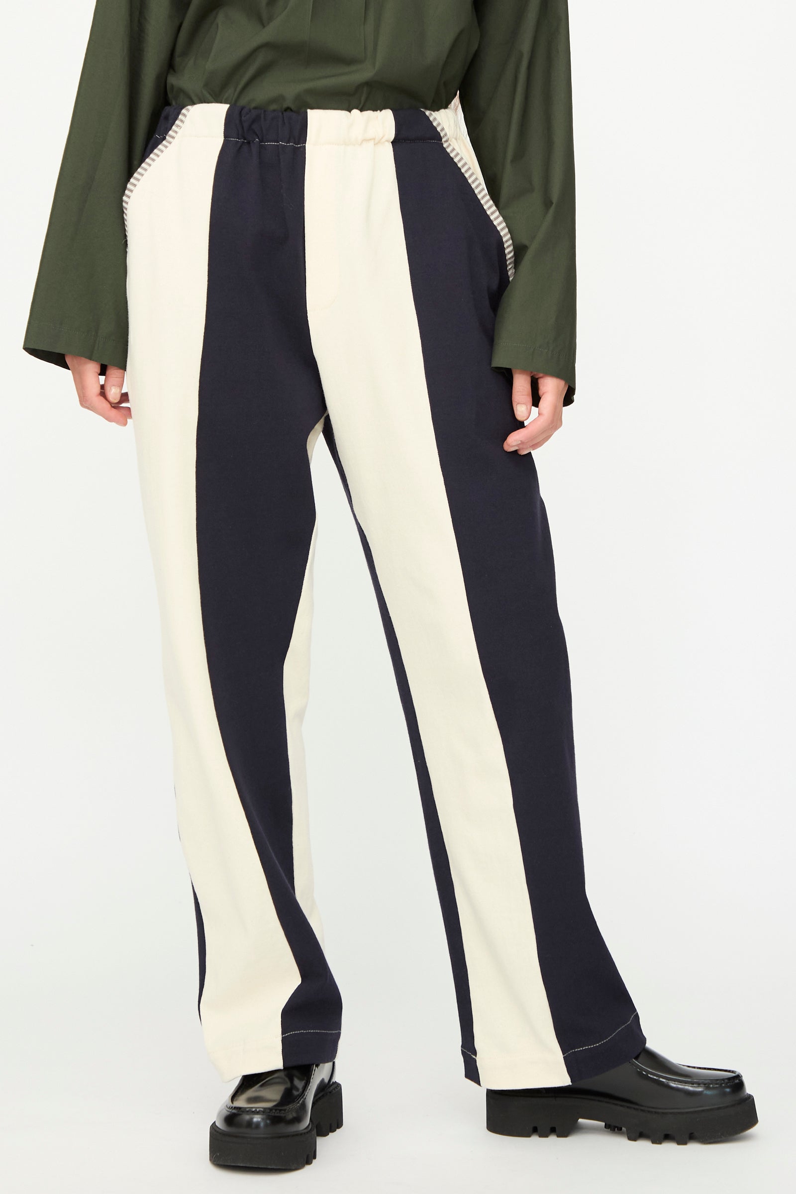 Wearing SC103's Rib Knit Leap Sweats in Index—black and white striped knit pants made in NYC—with a green long-sleeve top and black platform shoes, standing against a plain background.