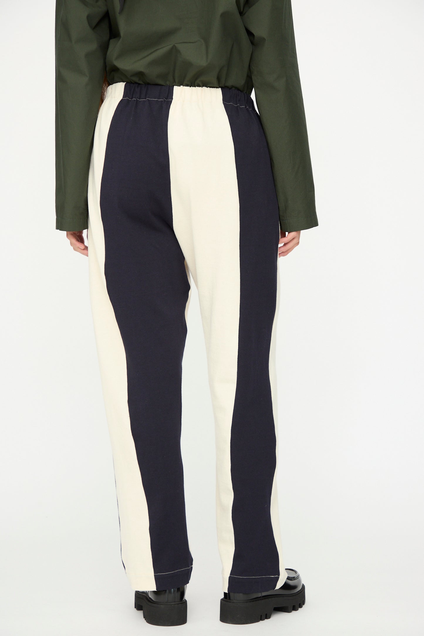 Person wearing SC103 Rib Knit Leap Sweats in Index—relaxed fit, high-waisted pants with wide black-and-white stripes—styled with dark shoes and a dark green long-sleeve shirt. Photographed from behind. Made in NYC.