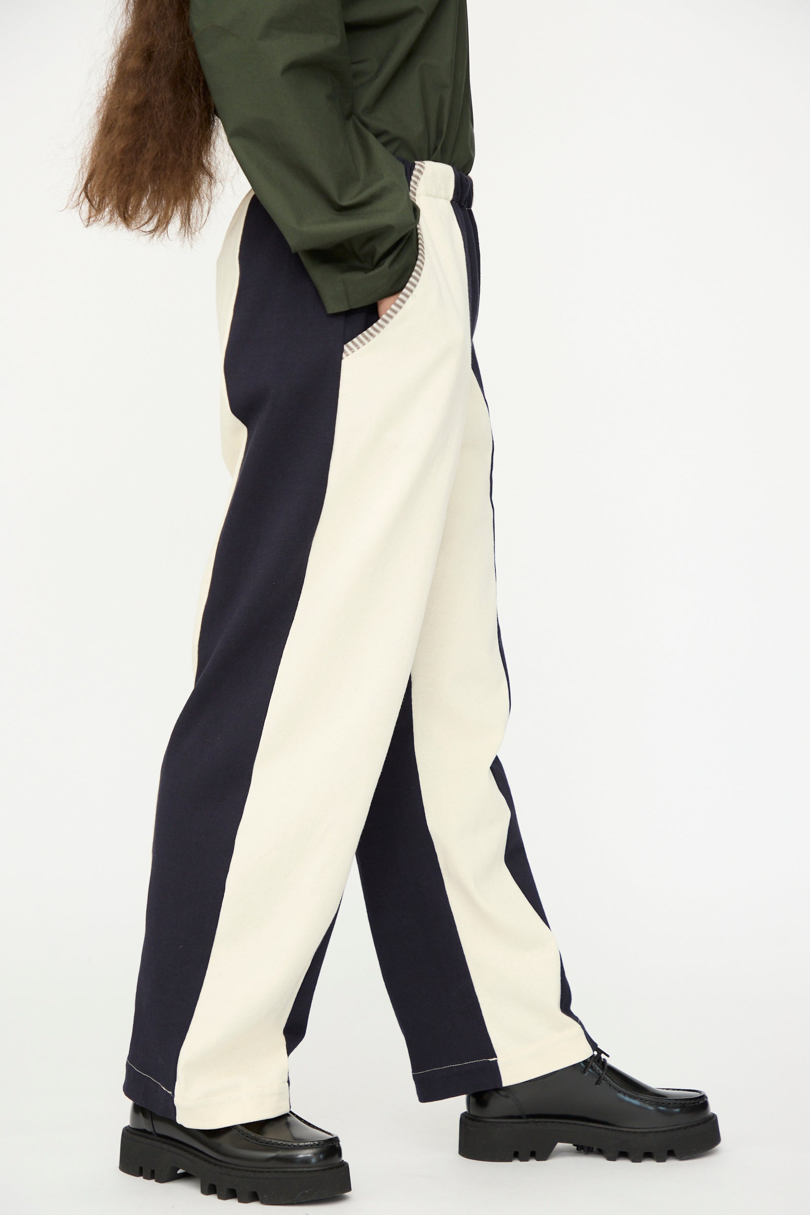 A person wearing SC103 Rib Knit Leap Sweats in Index—relaxed fit striped pants in black and white—with a dark green long-sleeve top and black platform shoes, shown from the side.