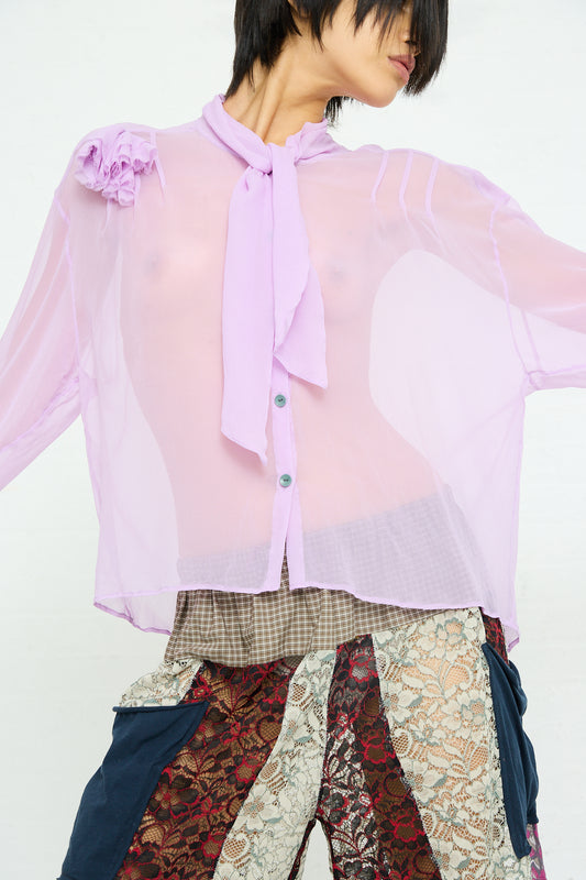 A person wearing a SC103 Silk Chiffon Mandolin Blouse in Sash with a rosette detail on the shoulder and patterned trousers.