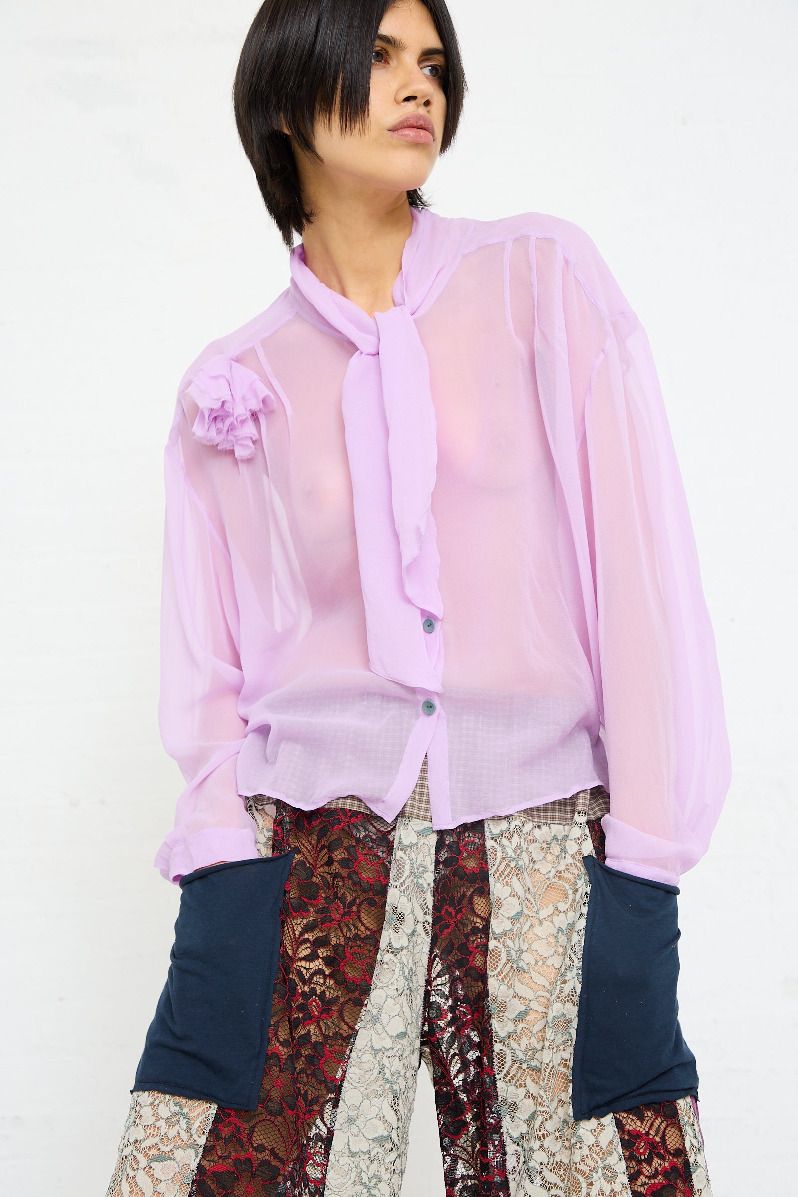 A person stands against a white background, wearing a SC103 Silk Chiffon Mandolin Blouse in Sash with rosette detail and patterned trousers.