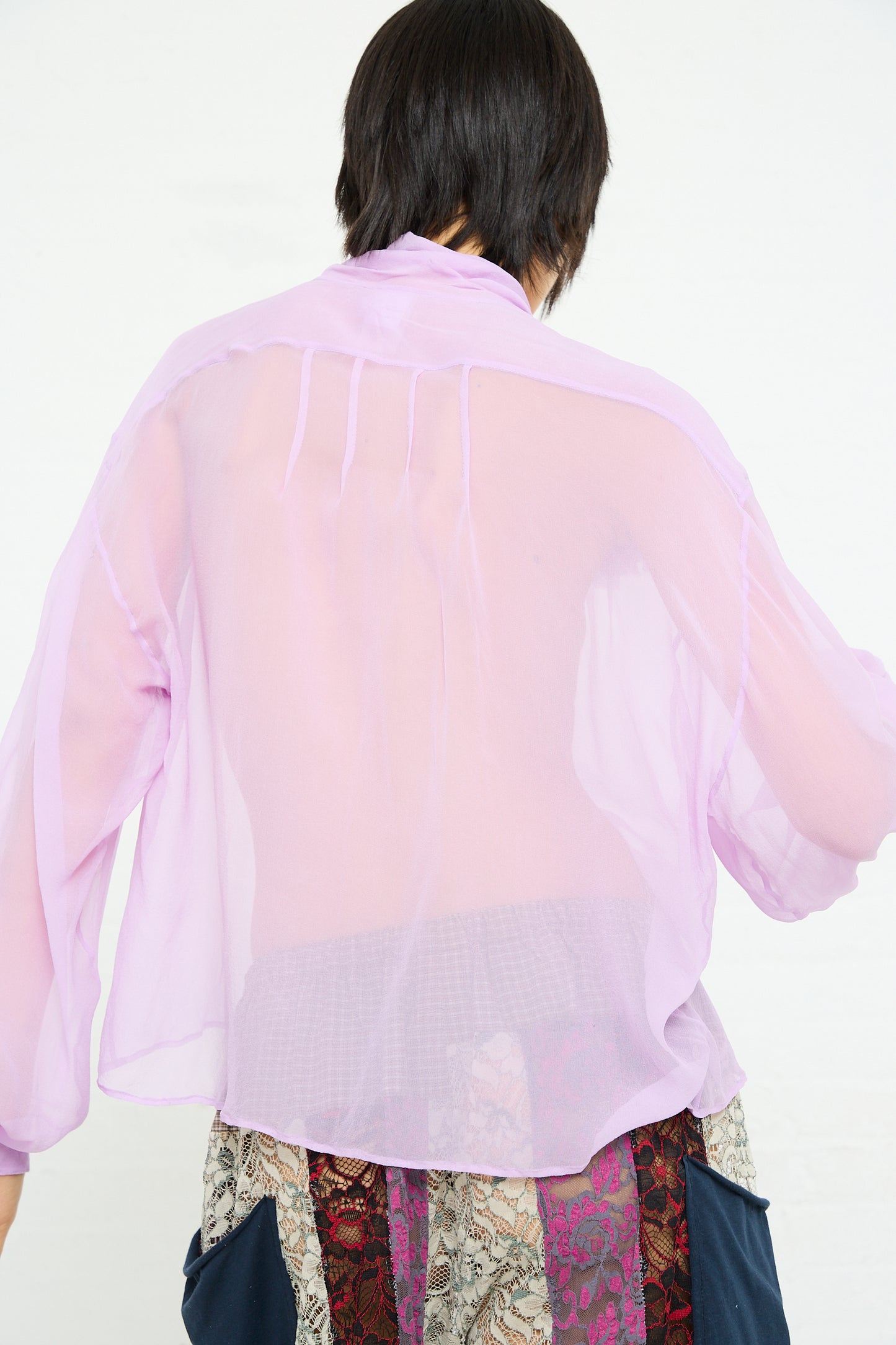 A person wearing a sheer pink Silk Chiffon Mandolin Blouse with rosette detail and patterned bottoms, viewed from behind.