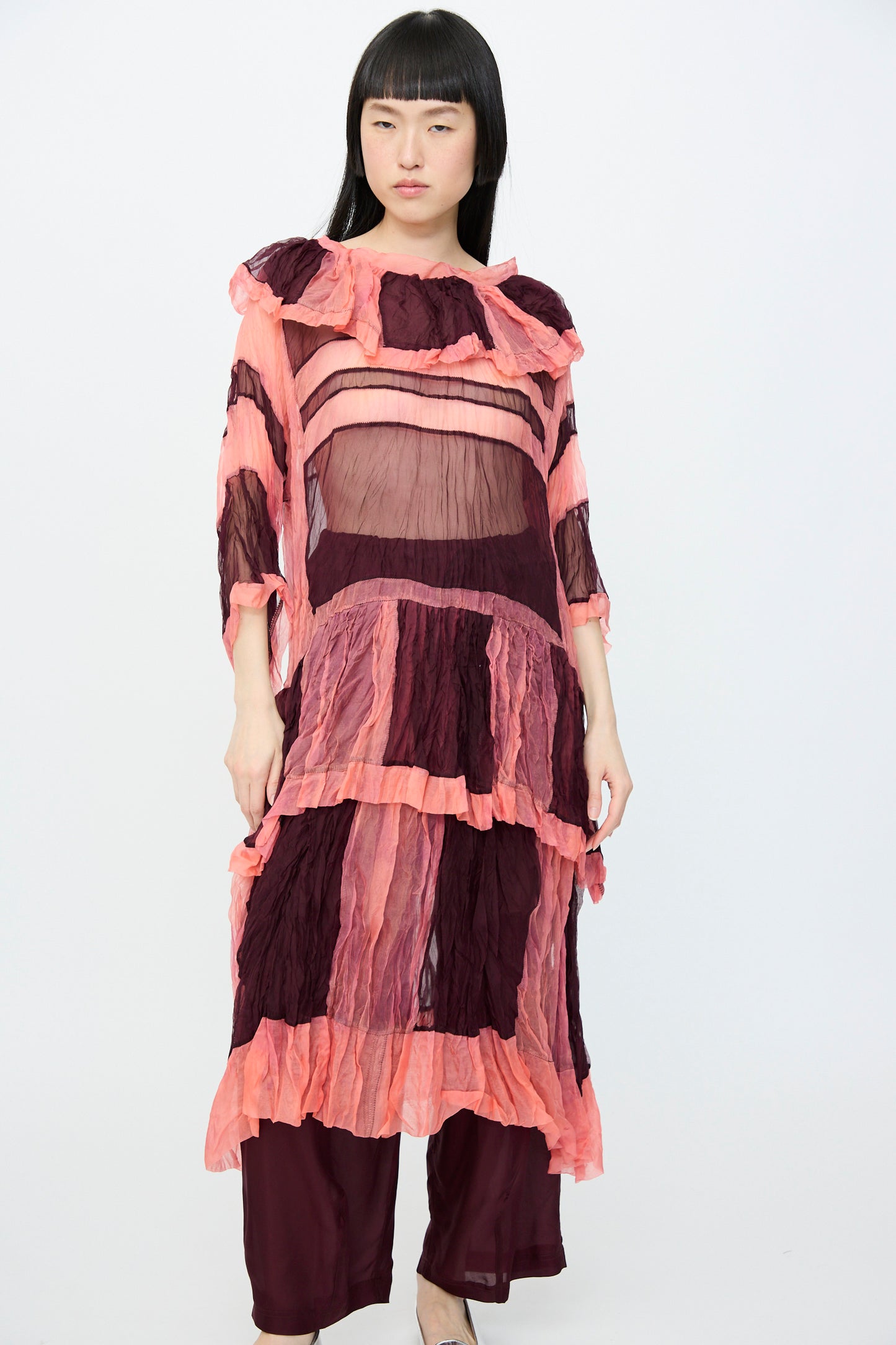A person stands against a plain background wearing SC103's oversized Silk Organza Ides Dress in Venus, featuring sheer pink and burgundy stripes over pants, with the silk organza layers.