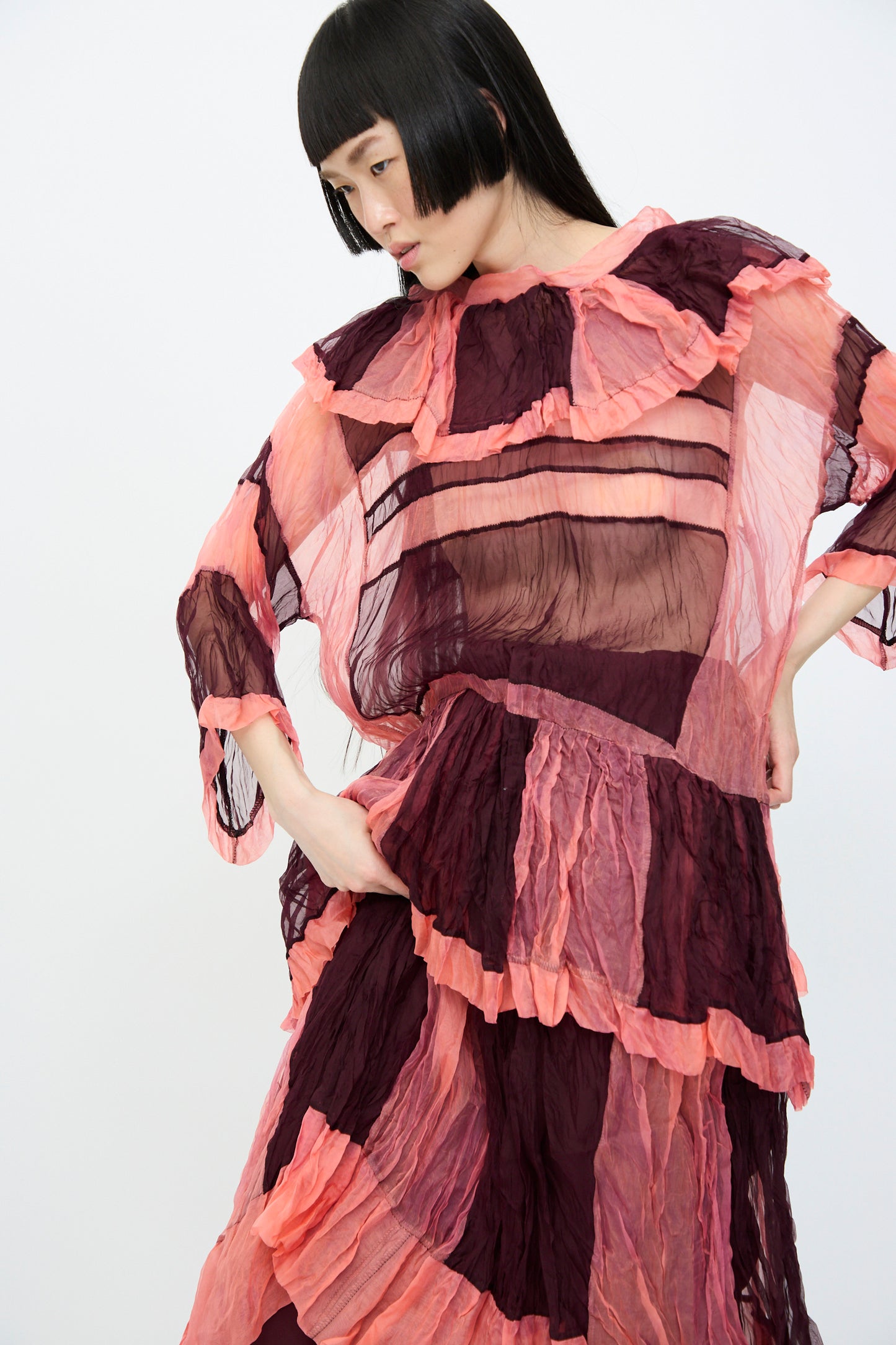 A person models the SC103 Silk Organza Ides Dress in Venus, a pink and maroon sheer ruffled midi dress with an oversized fit, against a plain white background, highlighting its layered design.