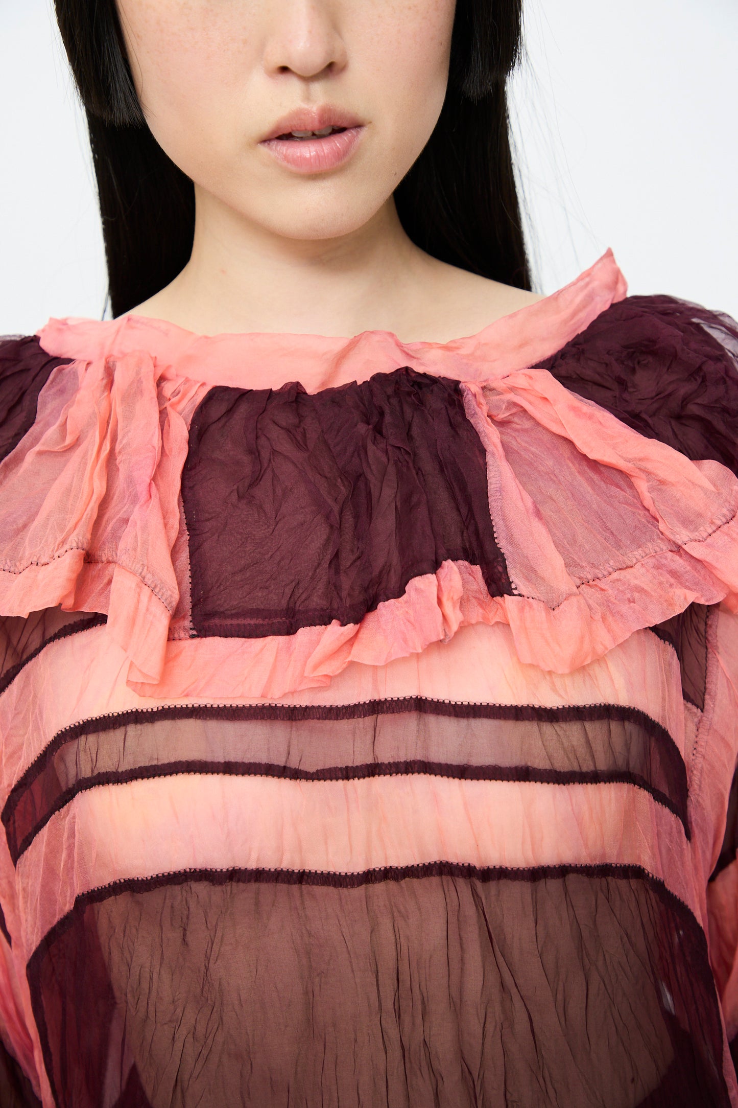 Close-up of a person wearing the SC103 Silk Organza Ides Dress in Venus, featuring sheer layers of pink and dark maroon silk organza. Only the lower face and neck are visible against a plain backdrop.