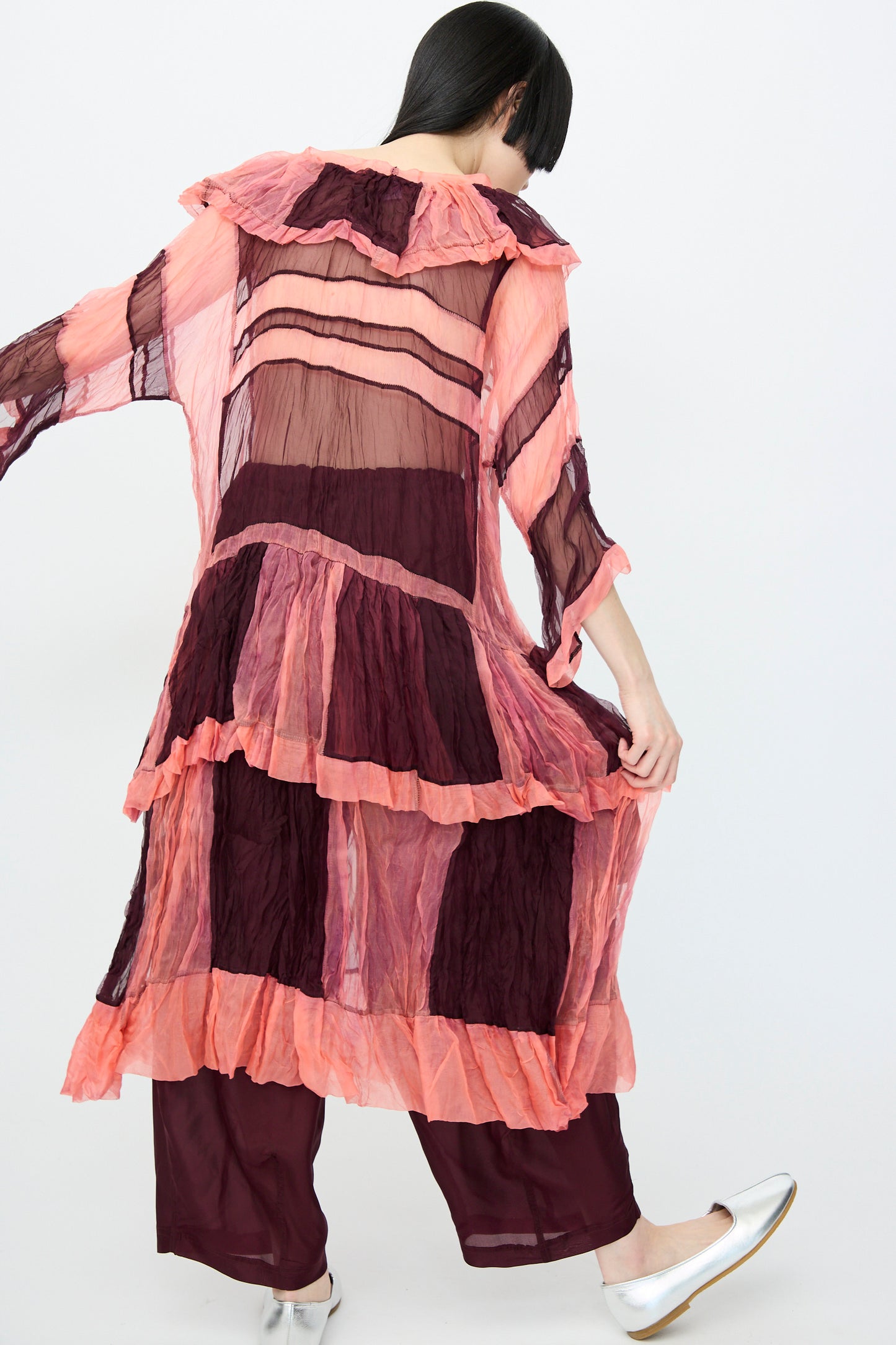 A person in a SC103 Silk Organza Ides Dress in Venus, featuring sheer layers and pink with burgundy stripes in an oversized fit, pairs it with burgundy pants and silver shoes against a plain backdrop.
