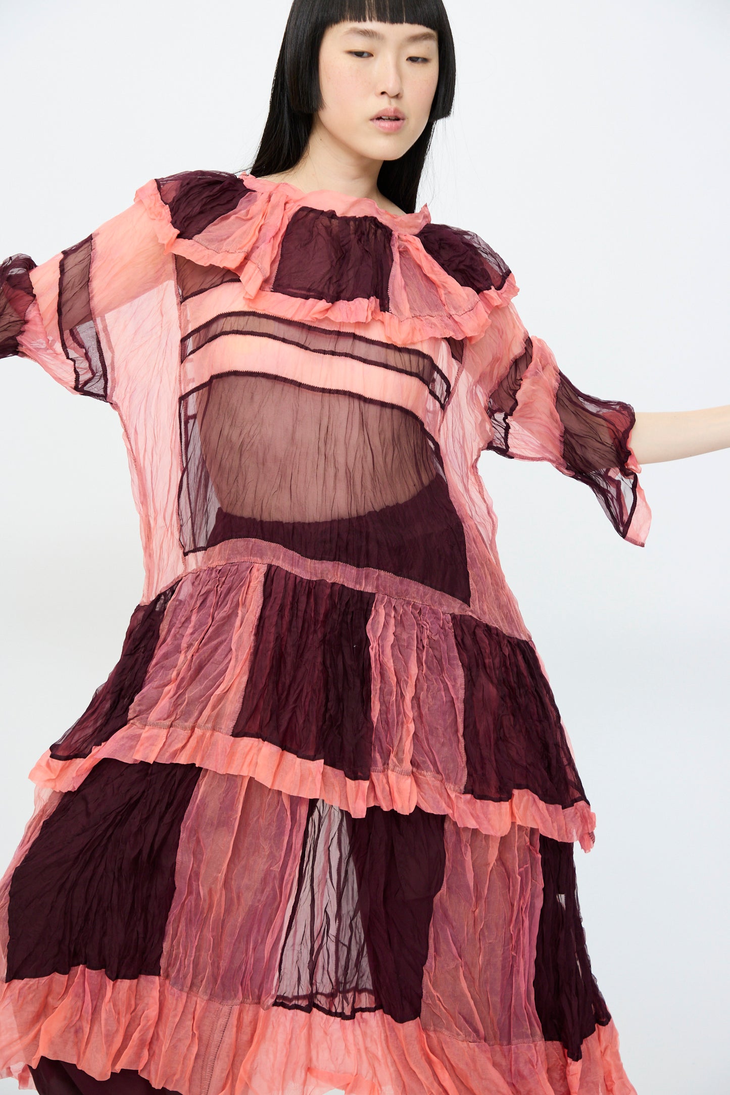 A model in the SC103 Silk Organza Ides Dress in Venus, featuring sheer pink and burgundy layers, stands against a white backdrop.