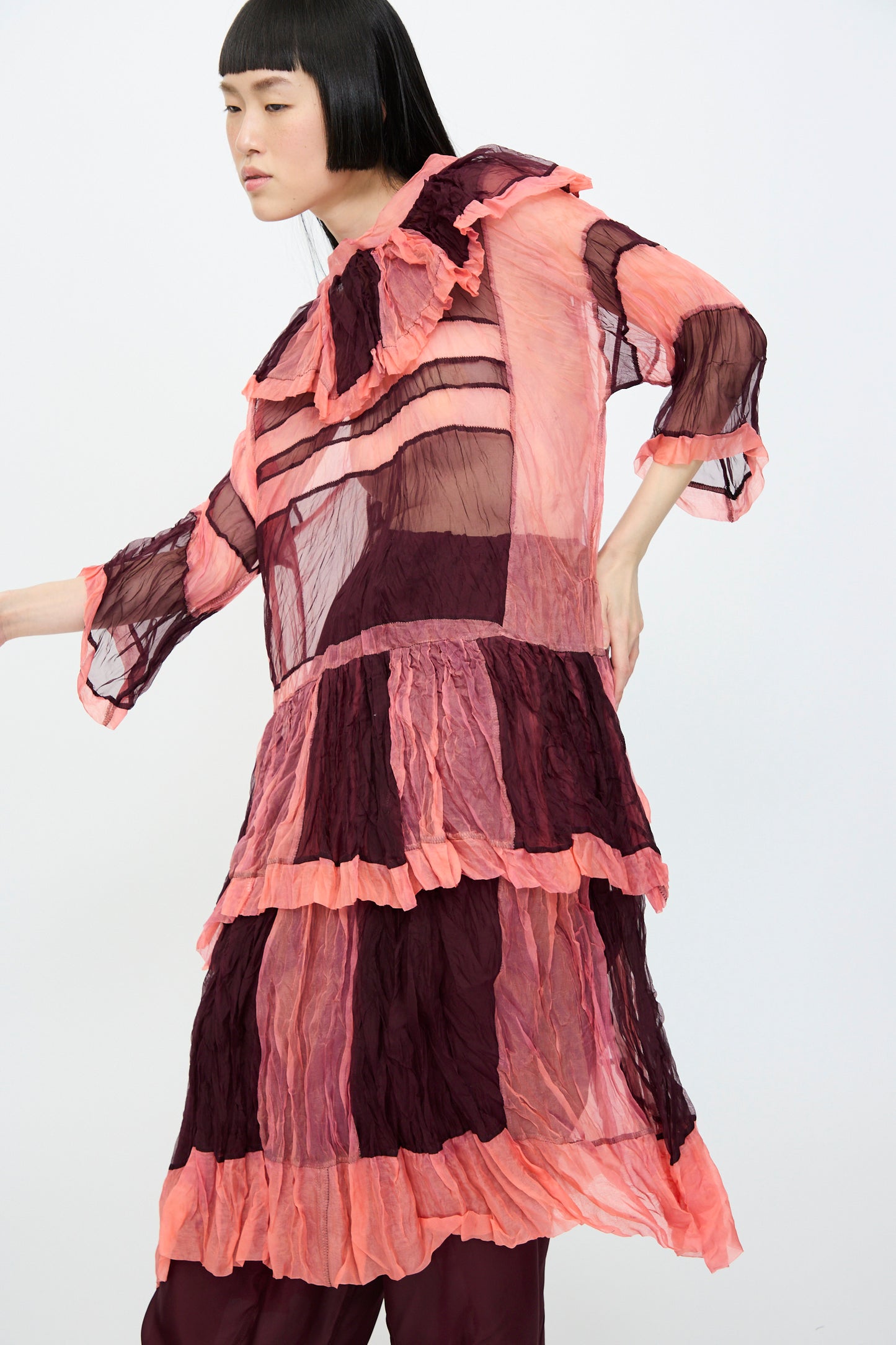 A person poses against a plain backdrop in the SC103 Silk Organza Ides Dress in Venus, a layered midi design made from sheer pink and burgundy silk organza.