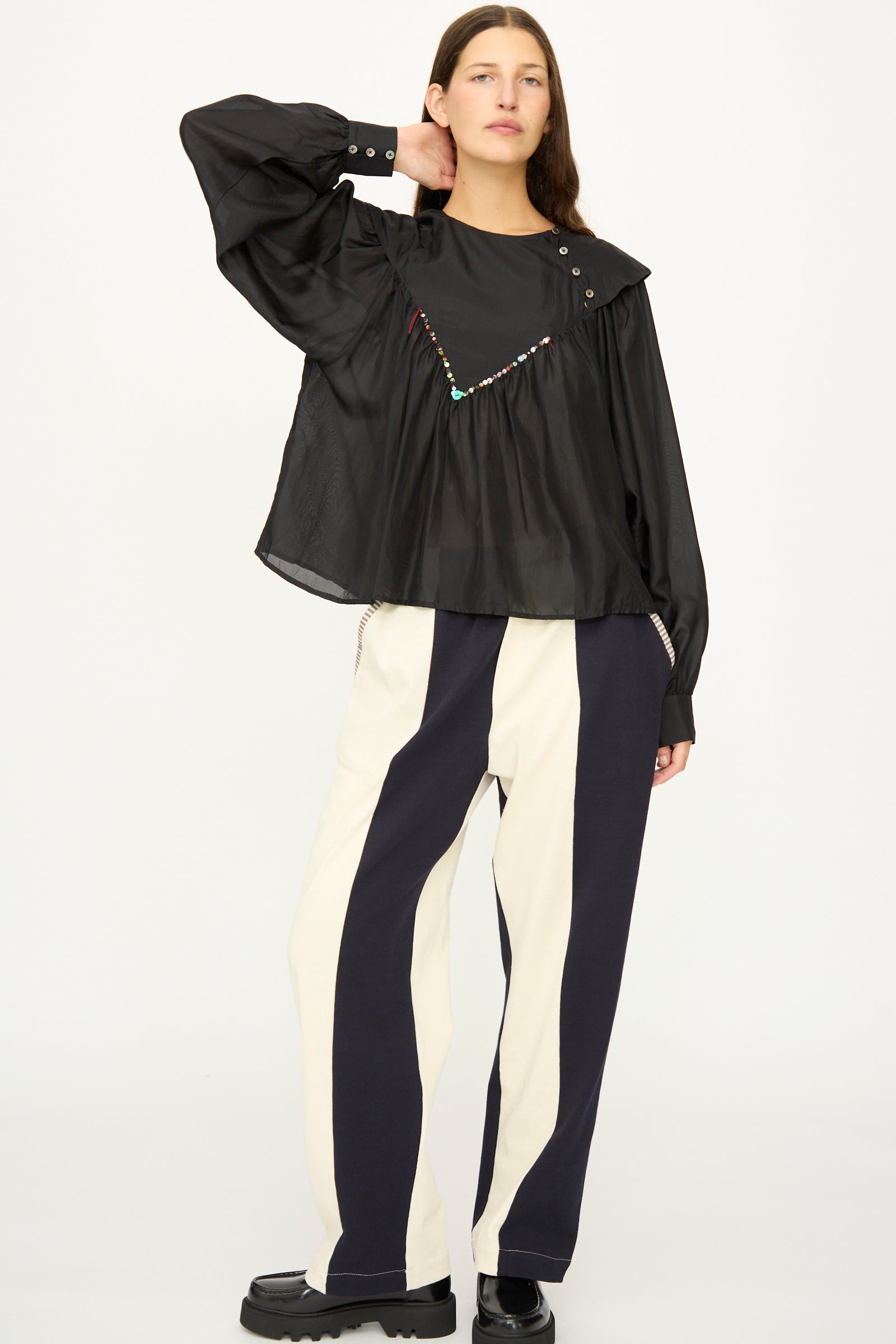 A person stands against a plain background wearing the SC103 Silk Prince Blouse in Thunder paired with wide-leg, high-contrast black and white striped pants and black shoes.