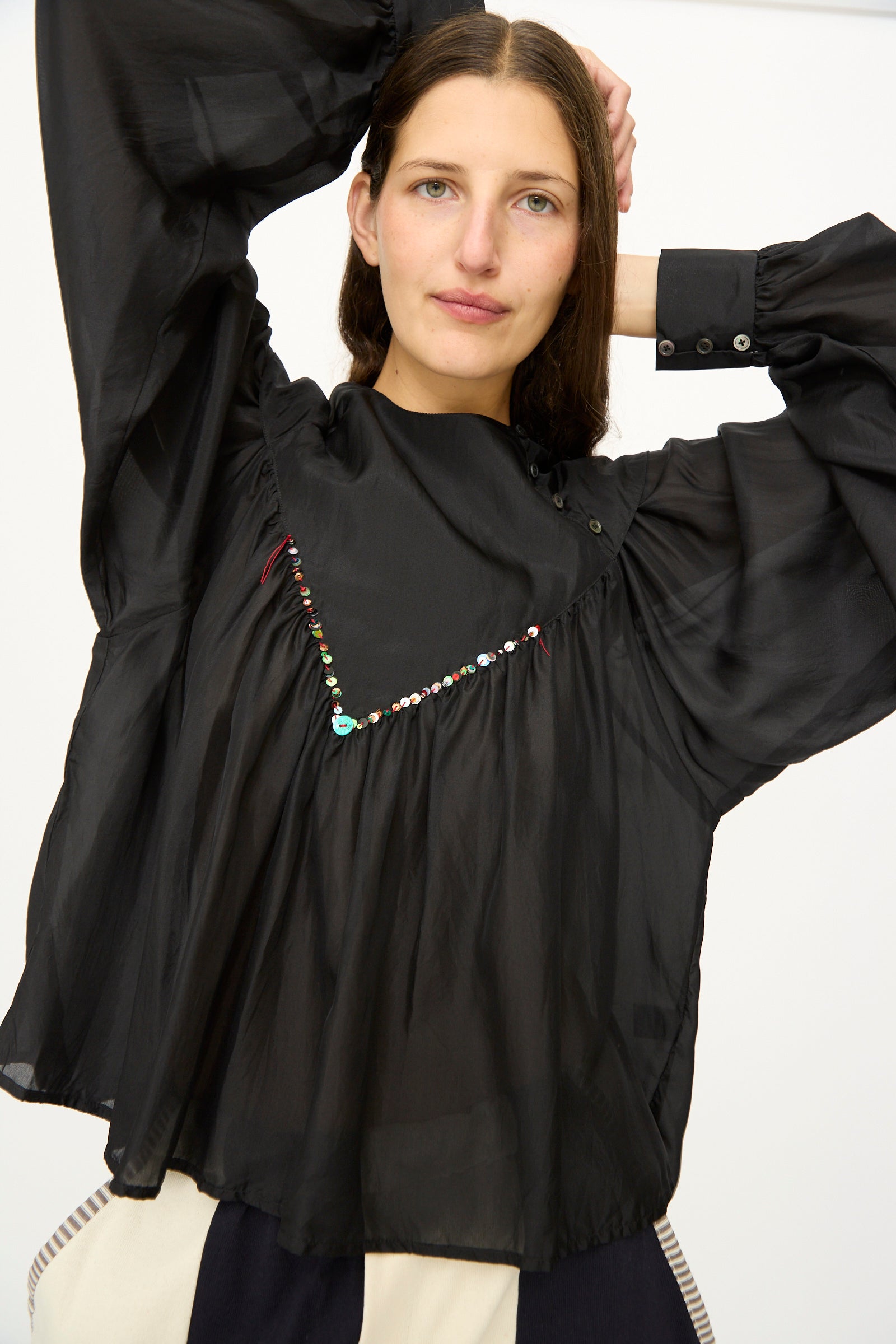 A woman stands with one arm raised, facing the camera, wearing the SC103 Silk Prince Blouse in Thunder— a loose, sheer black long sleeve top featuring sequin details.