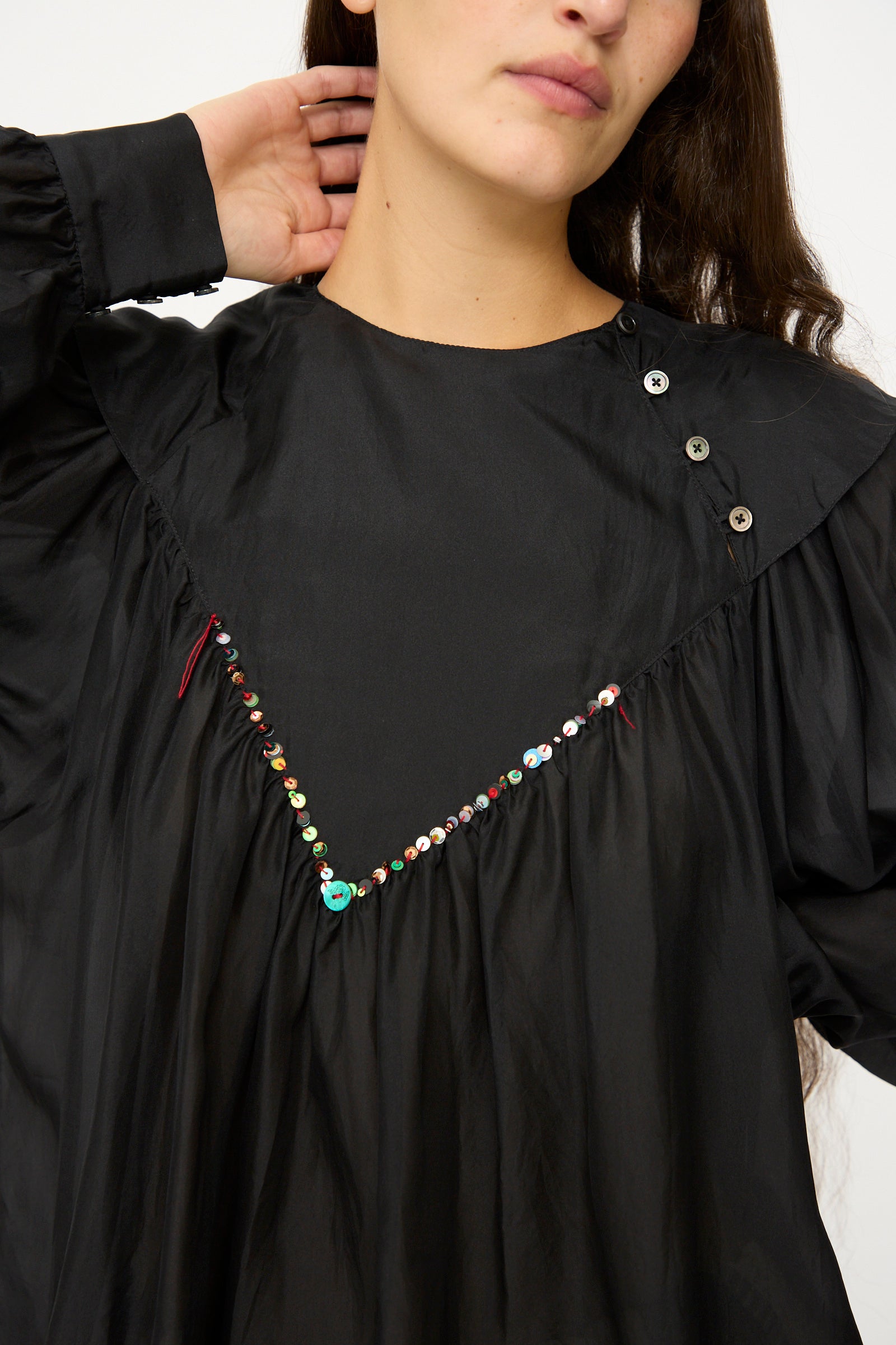 A woman wears the SC103 Silk Prince Blouse in Thunder, a long sleeve top with a V-shaped line of embroidered sequins and buttons near the shoulder, one hand gently touching her neck.