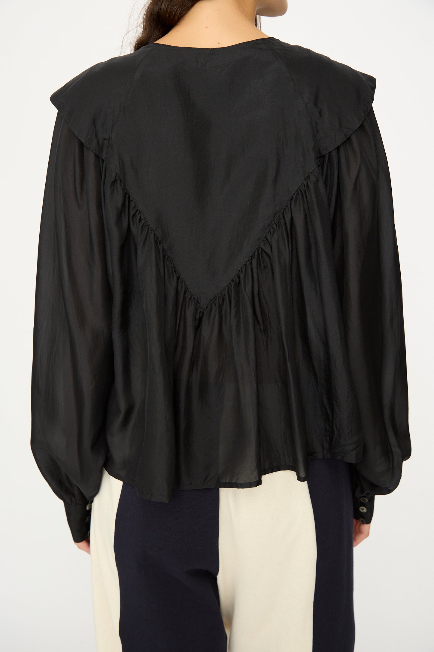A person wearing the SC103 Silk Prince Blouse in Thunder, a loose-fitting black top with a triangular yoke detail on the back, paired with black-and-white pants, photographed from behind.