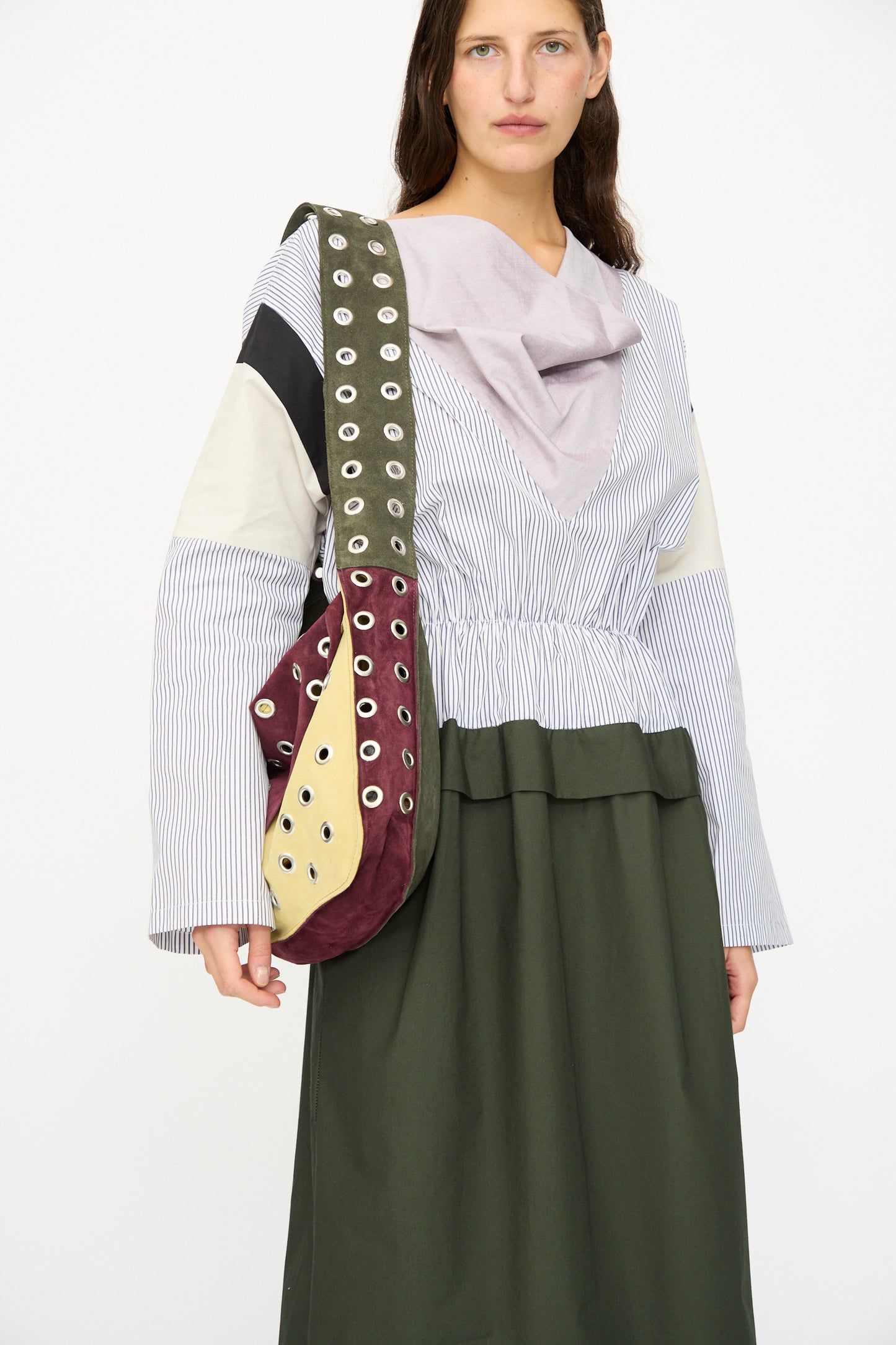 A woman poses against a white background wearing a long-sleeve, multi-patterned dress and the SC103 Suede Record Messenger in Campus crossbody bag.