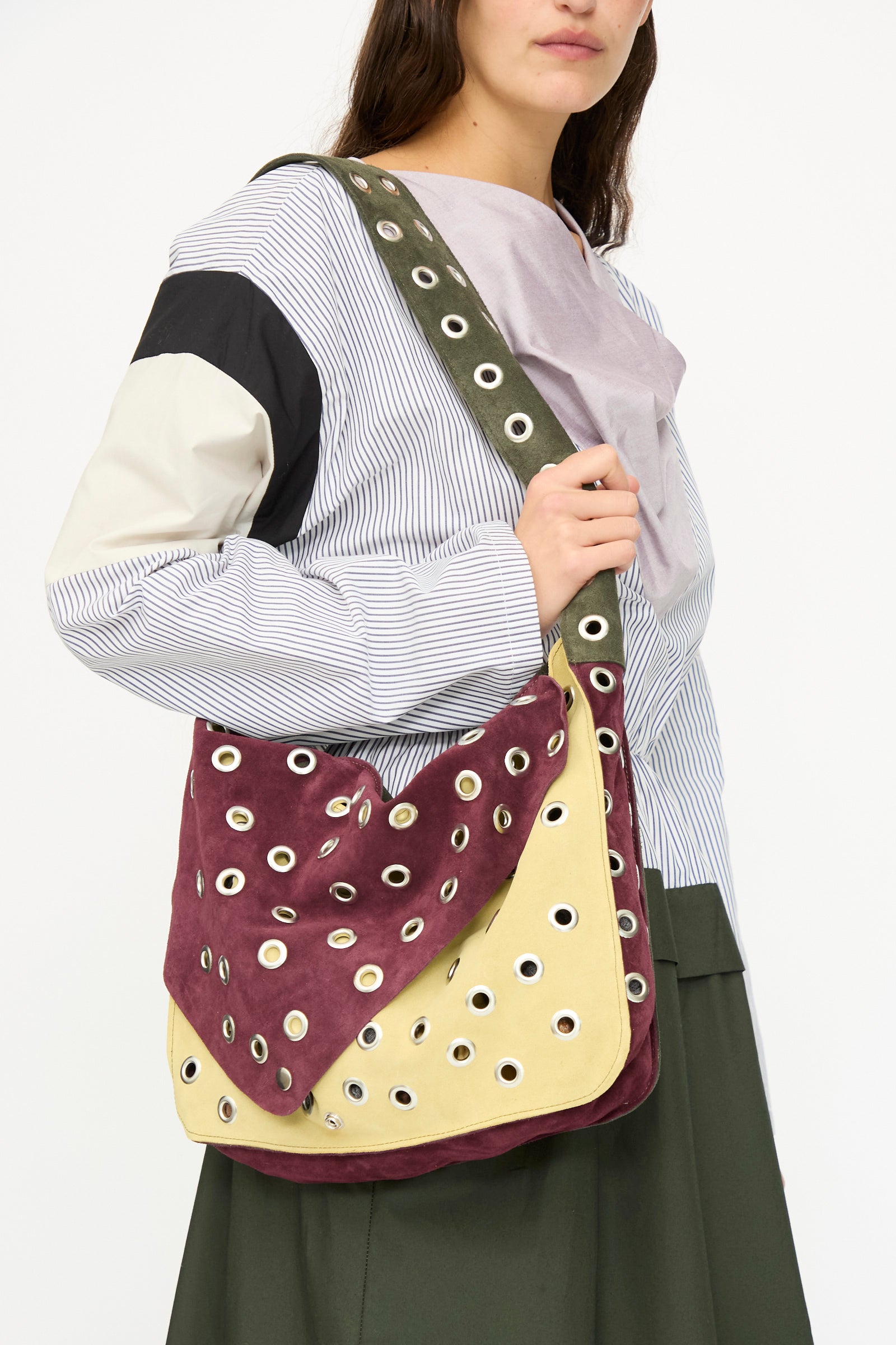 A woman in a multicolored shirt and dark skirt carries the SC103 Suede Record Messenger in Campus, a large two-tone patchwork bag with eyelet embellishments and an olive green strap.