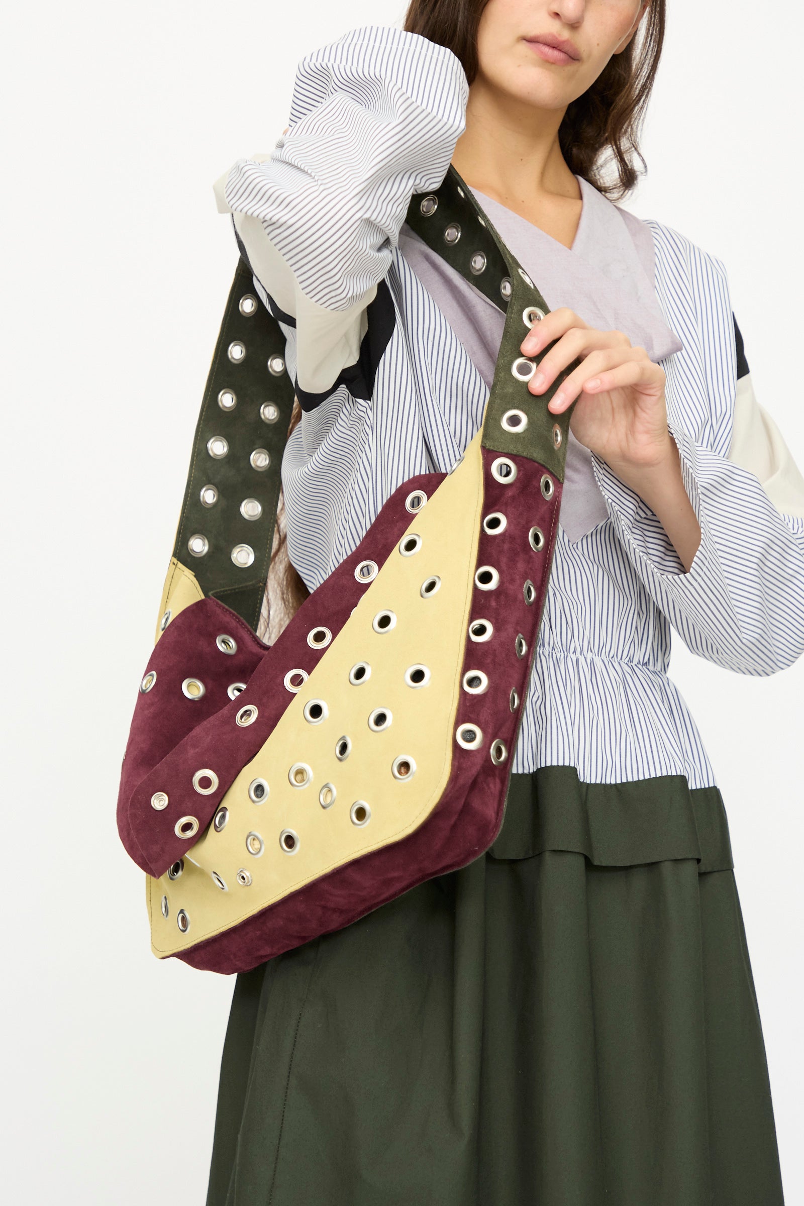 A woman in a striped blouse and green skirt carries the SC103 Suede Record Messenger in Campus, a large patchwork bag with maroon, yellow, and green panels, accented with silver grommets.
