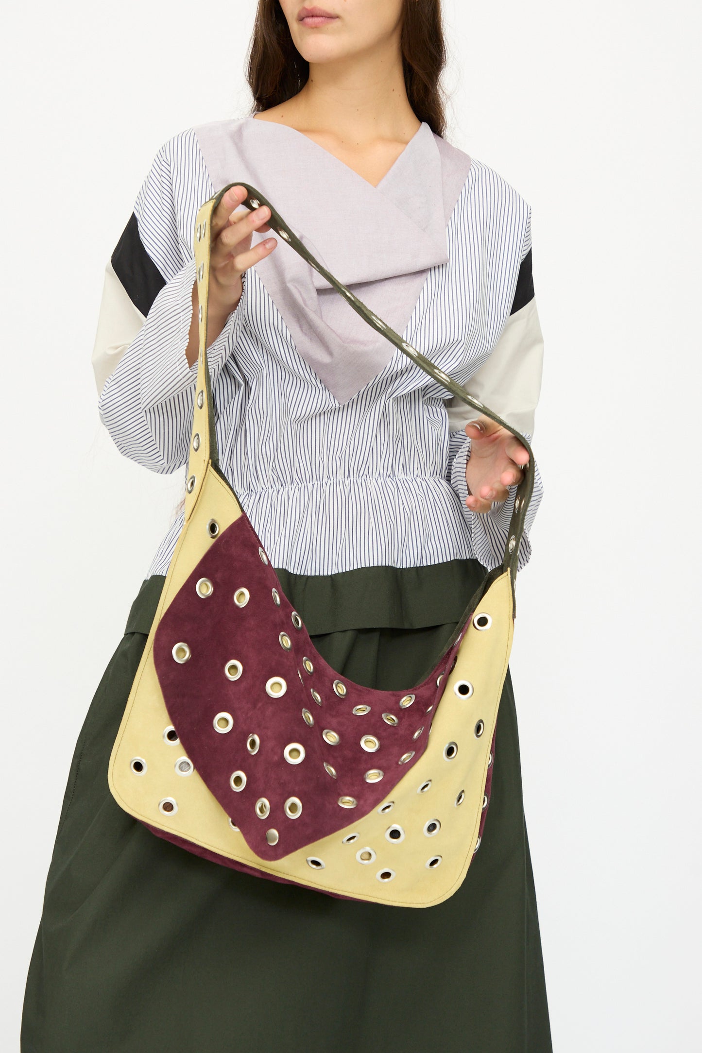 A woman holds the SC103 Suede Record Messenger in Campus, a large two-toned suede patchwork shoulder bag with metal eyelet details. She pairs it with a long-sleeved, multi-patterned blouse and a dark skirt.