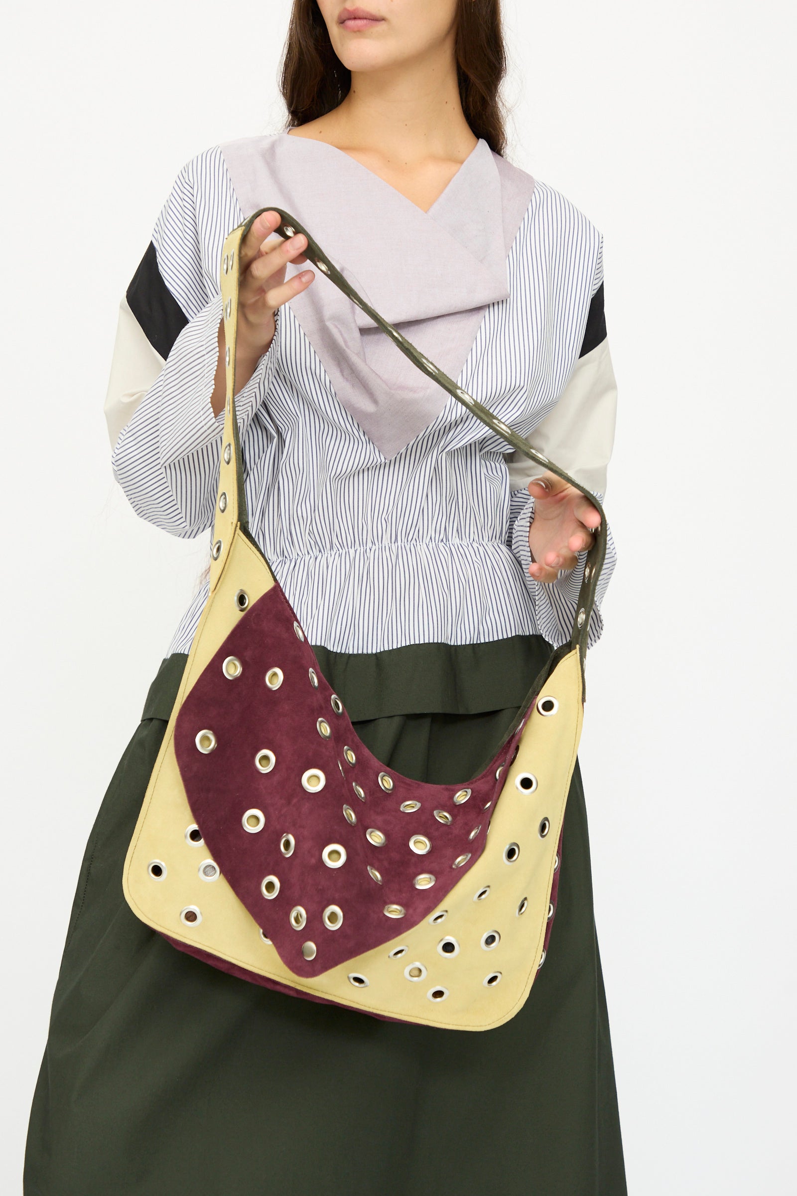 A woman holds the SC103 Suede Record Messenger in Campus, a large two-toned suede patchwork shoulder bag with metal eyelet details. She pairs it with a long-sleeved, multi-patterned blouse and a dark skirt.
