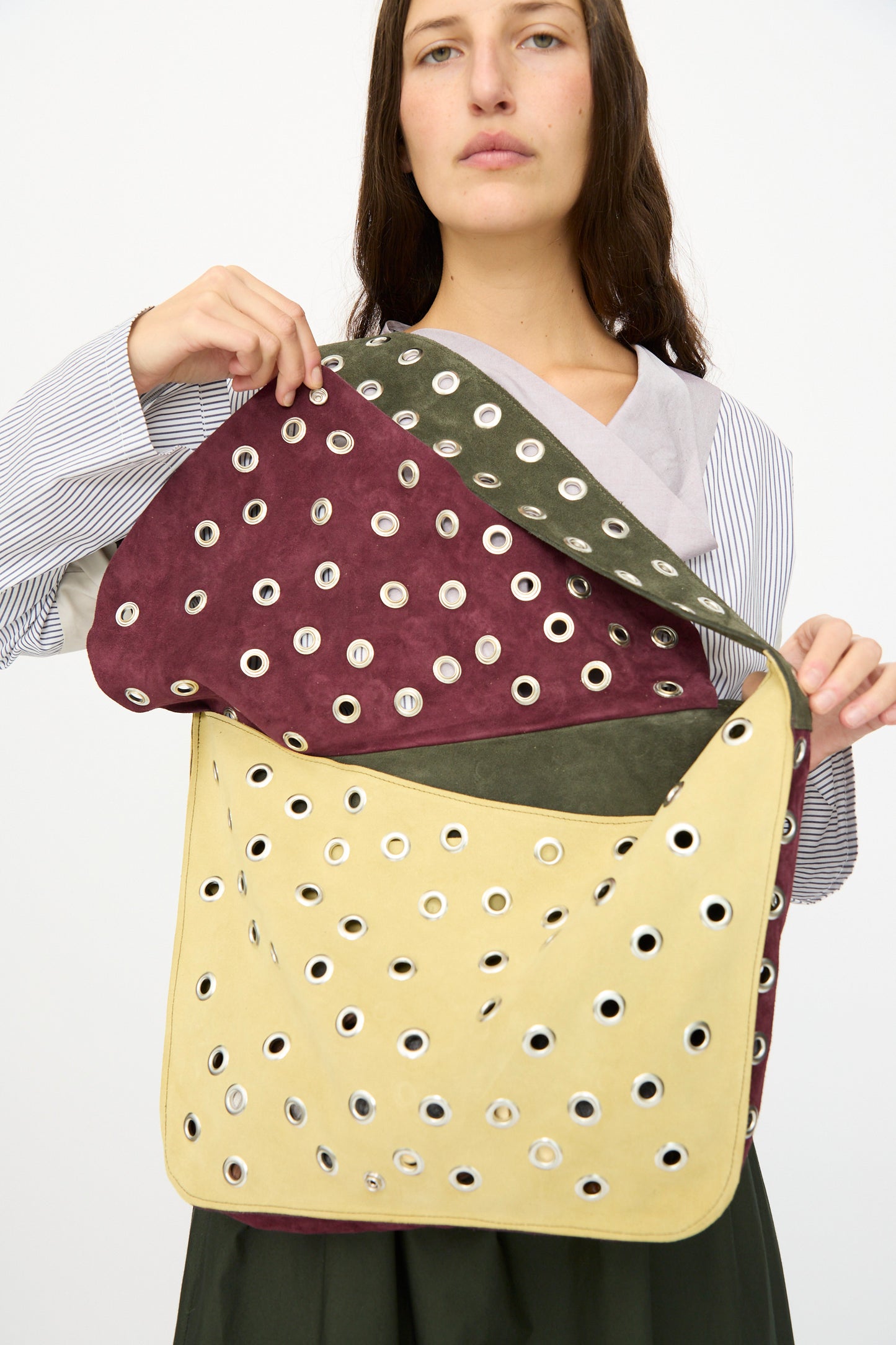 A person holds the SC103 Suede Record Messenger in Campus, an oversized bag with silver grommet holes, partially open to showcase its spacious interior and distinctive multicolored suede patchwork design.