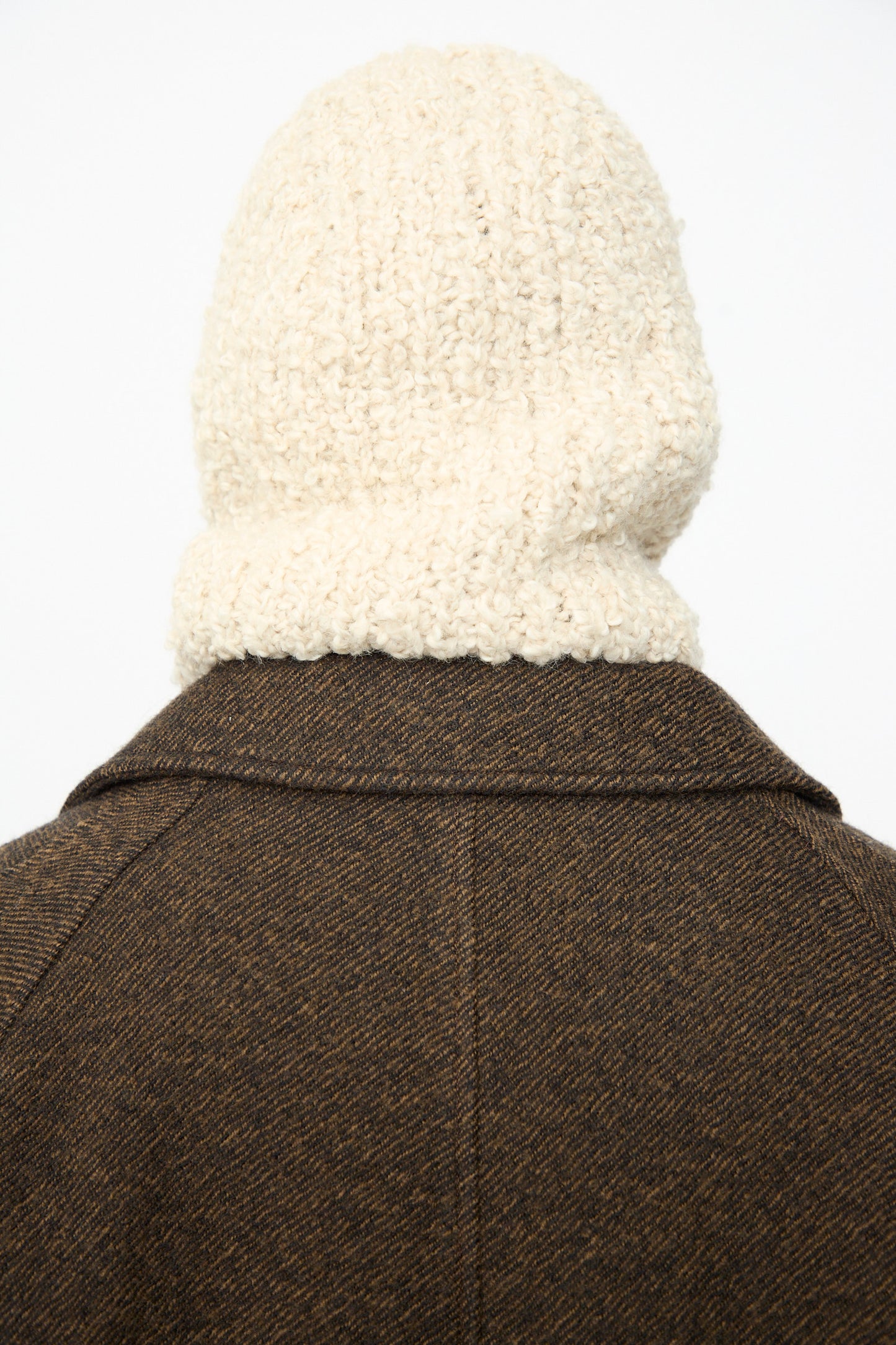 A person wearing a brown wool alpaca blend coat and the SONO Bruna Balaclava in Natural, featuring a thick, cream-colored knit with a subtle ribbed texture, photographed from behind against a plain light background.