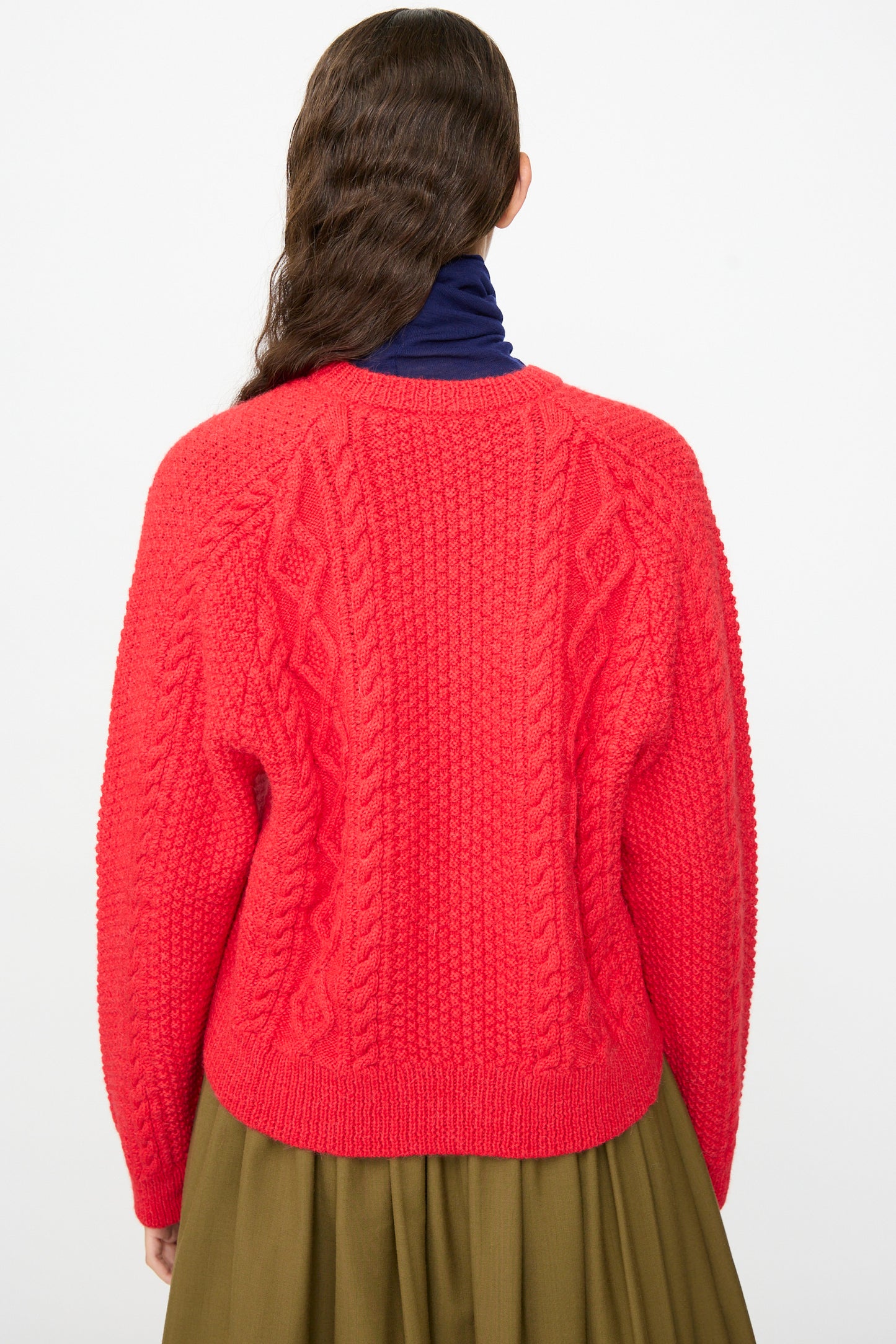 Person with long wavy brown hair, shown from the back against a plain background, wearing the SONO Candy Cardigan in Red—a textured wool alpaca cardigan made in the UK—layered over a dark blue turtleneck and an olive green skirt.