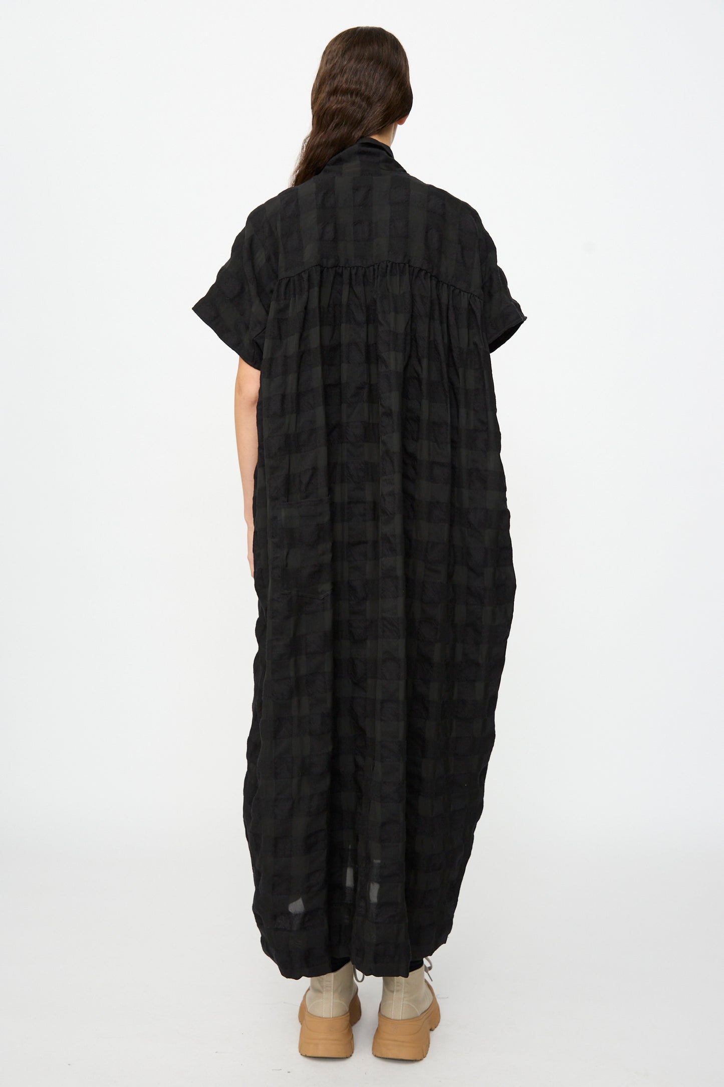 A person stands with their back to the camera, wearing the SONO Dawn Dress in Black on Black—a long, loose-fitting checkered maxi dress crafted from a wool-viscose blend—paired with beige shoes, set against a plain white background.