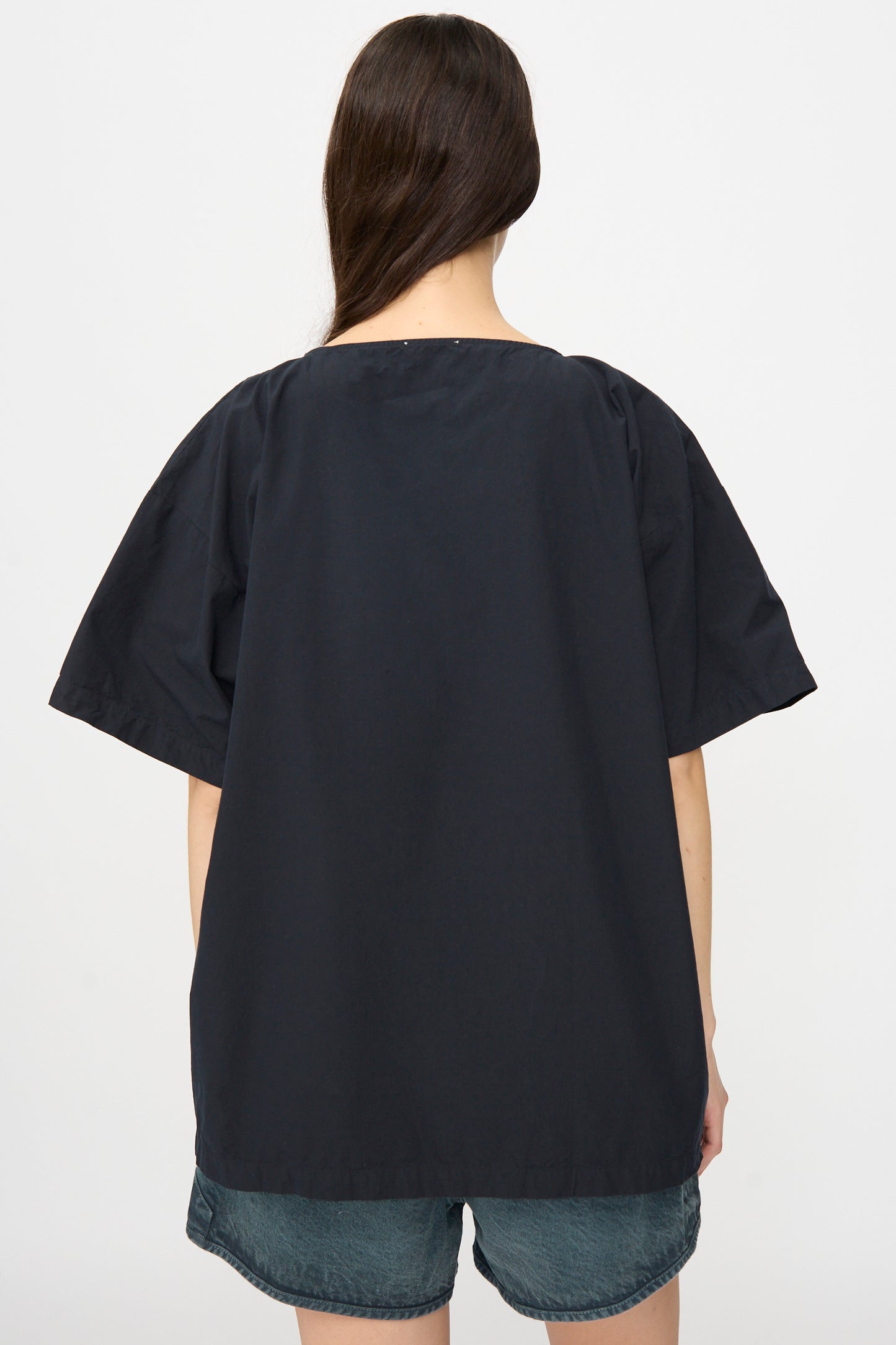 A person with long brown hair is shown from the back against a plain light background, wearing the SONO Friedrich Tee in Off Black and denim shorts.