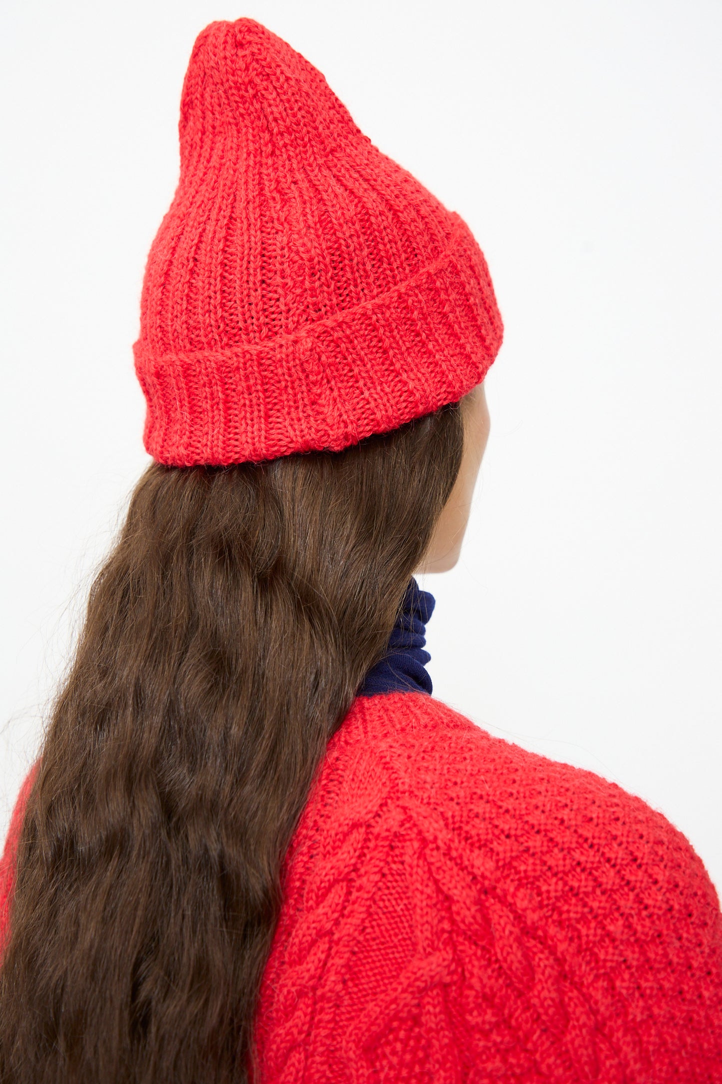 A person with long wavy brown hair is shown from behind against a plain white background, wearing the SONO Gyu Beanie in Red and a matching red sweater.