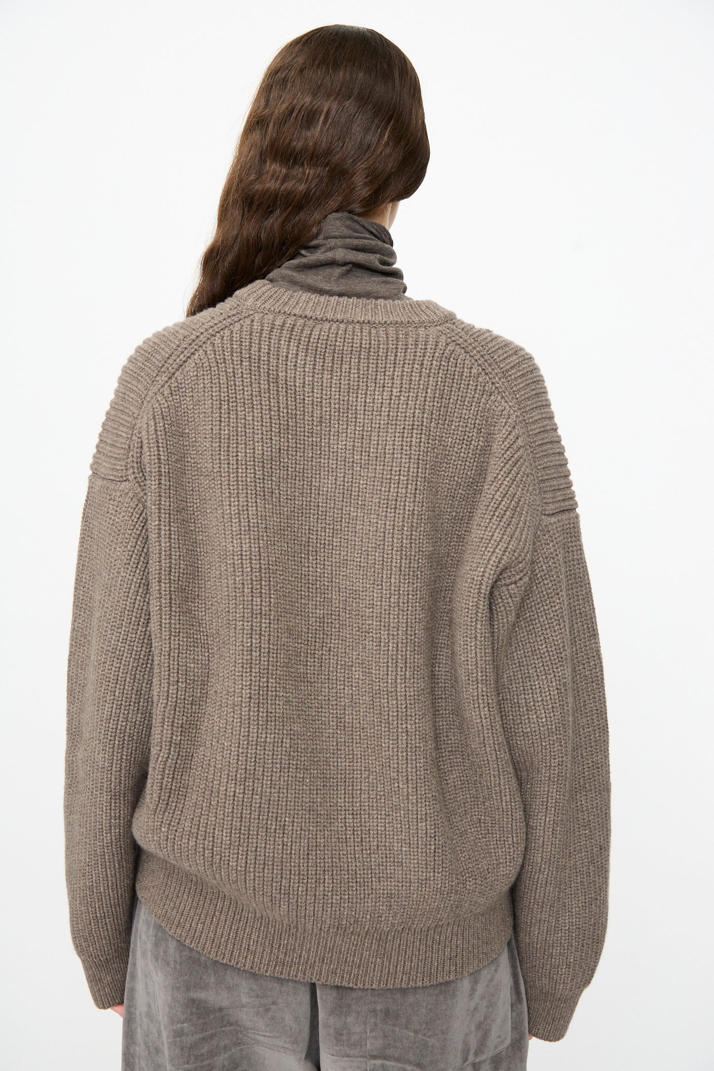 A person with long brown hair stands facing away against a plain white background, wearing the Joel Jumper in Smoke by SONO—a thick, ribbed knit wool taupe sweater—layered over a gray turtleneck top and paired with gray velvety pants.