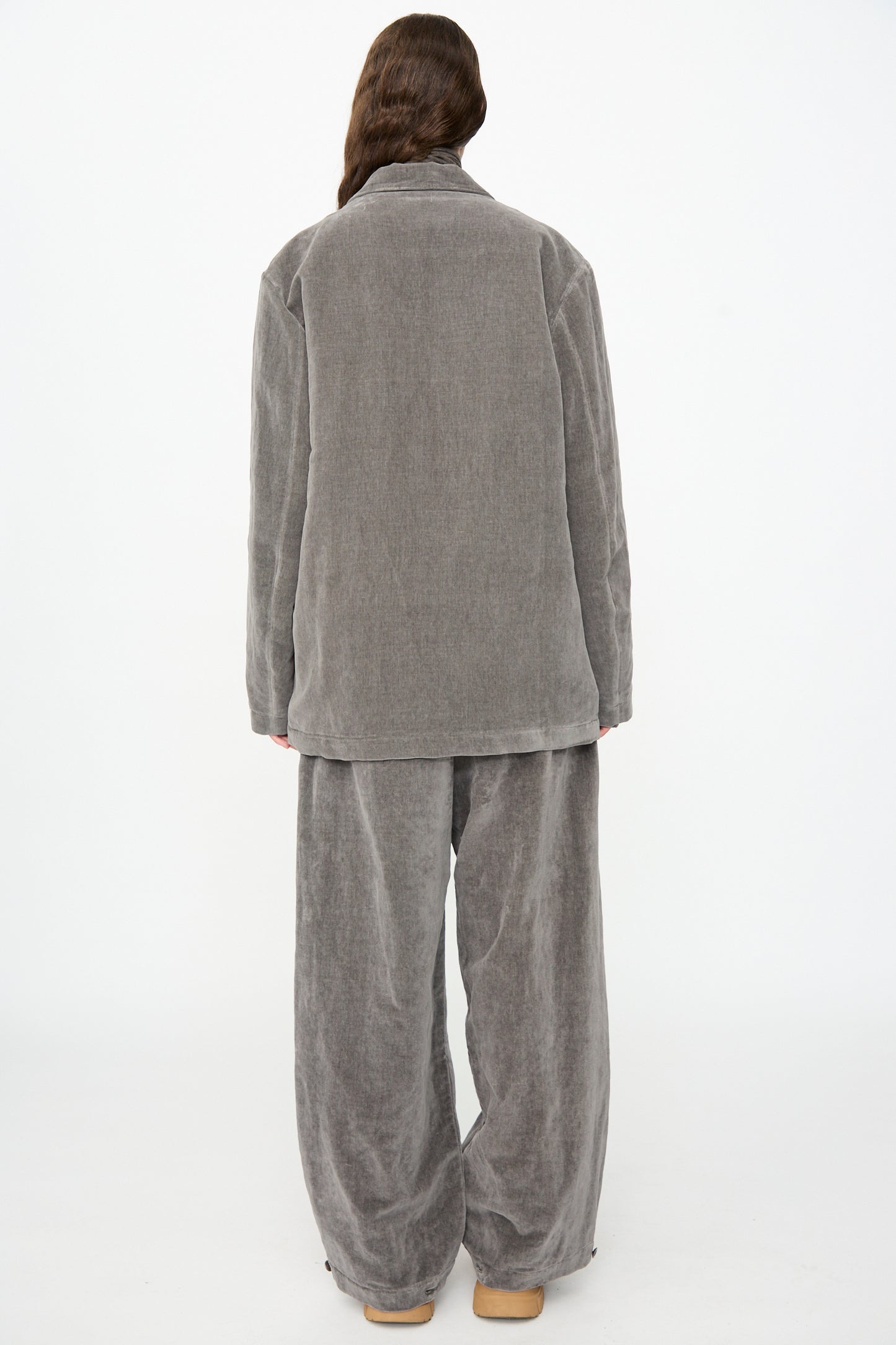 A person stands facing away, wearing the SONO Juri Jacket in Elephant—a loose-fitting, light gray corduroy suit with an oversized jacket and wide-leg pants—paired with flat tan shoes, set against a plain white background.