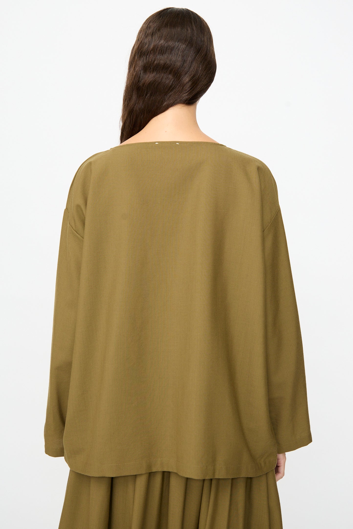A person with long brown hair is photographed from the back against a plain white background, wearing the SONO Laik Longsleeve in Kelp—an olive-green, oversized fit long-sleeve top paired with a matching skirt.